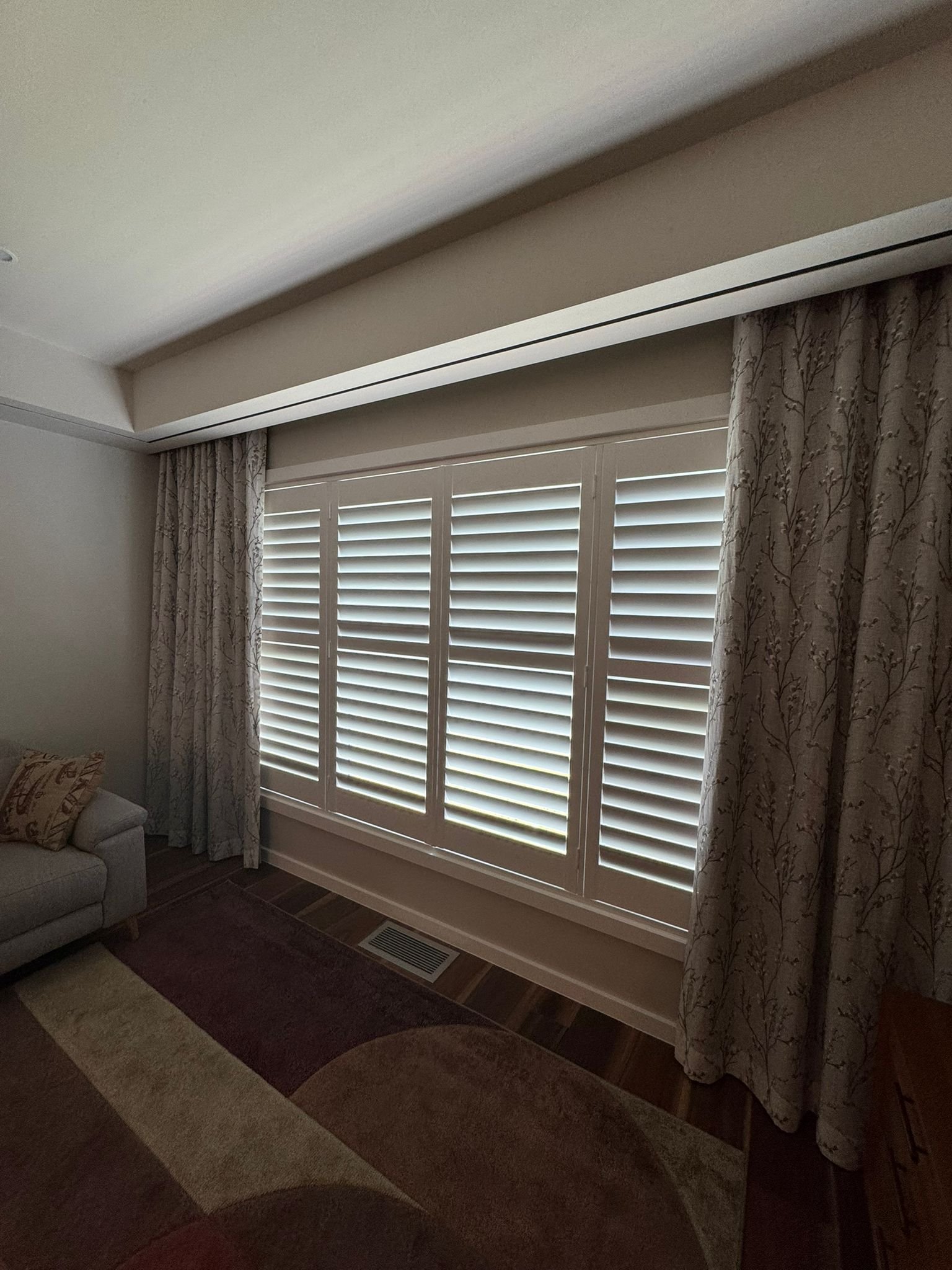 A room with beige walls, a large window with white plantation shutters, and curtains with a floral pattern. There is a small sofa with decorative pillows, a rug with a geometric pattern, and a floor vent visible.