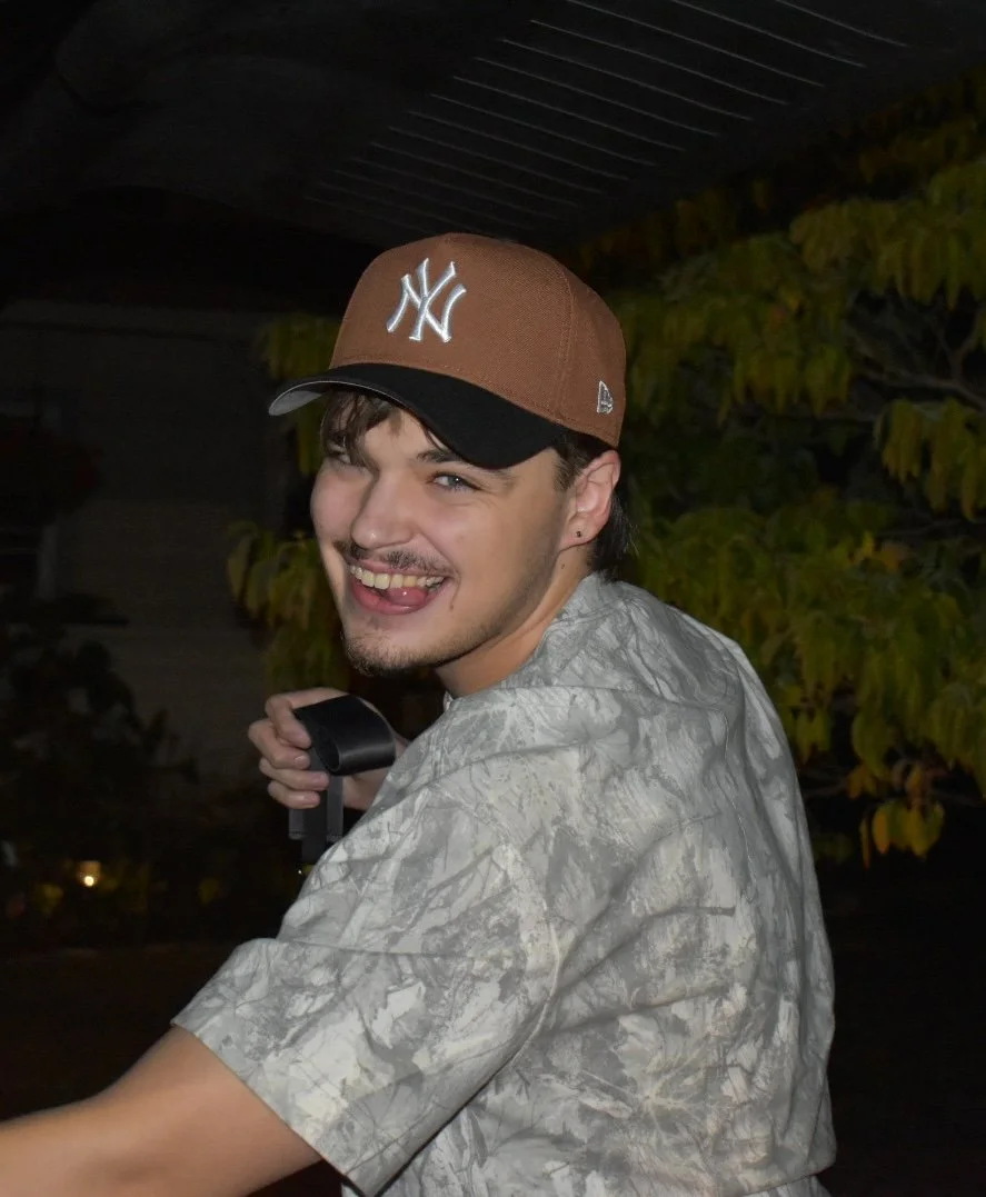 Man wearing a camouflage shirt sticking his tongue out wearing a brown yankees hat