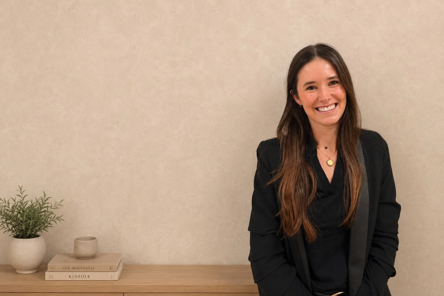 A smiling young woman with long brown hair, wearing a black blazer and a gold necklace, stands in front of a beige wall. A wooden shelf with a small plant, a candle holder, and two books is visible to her left.