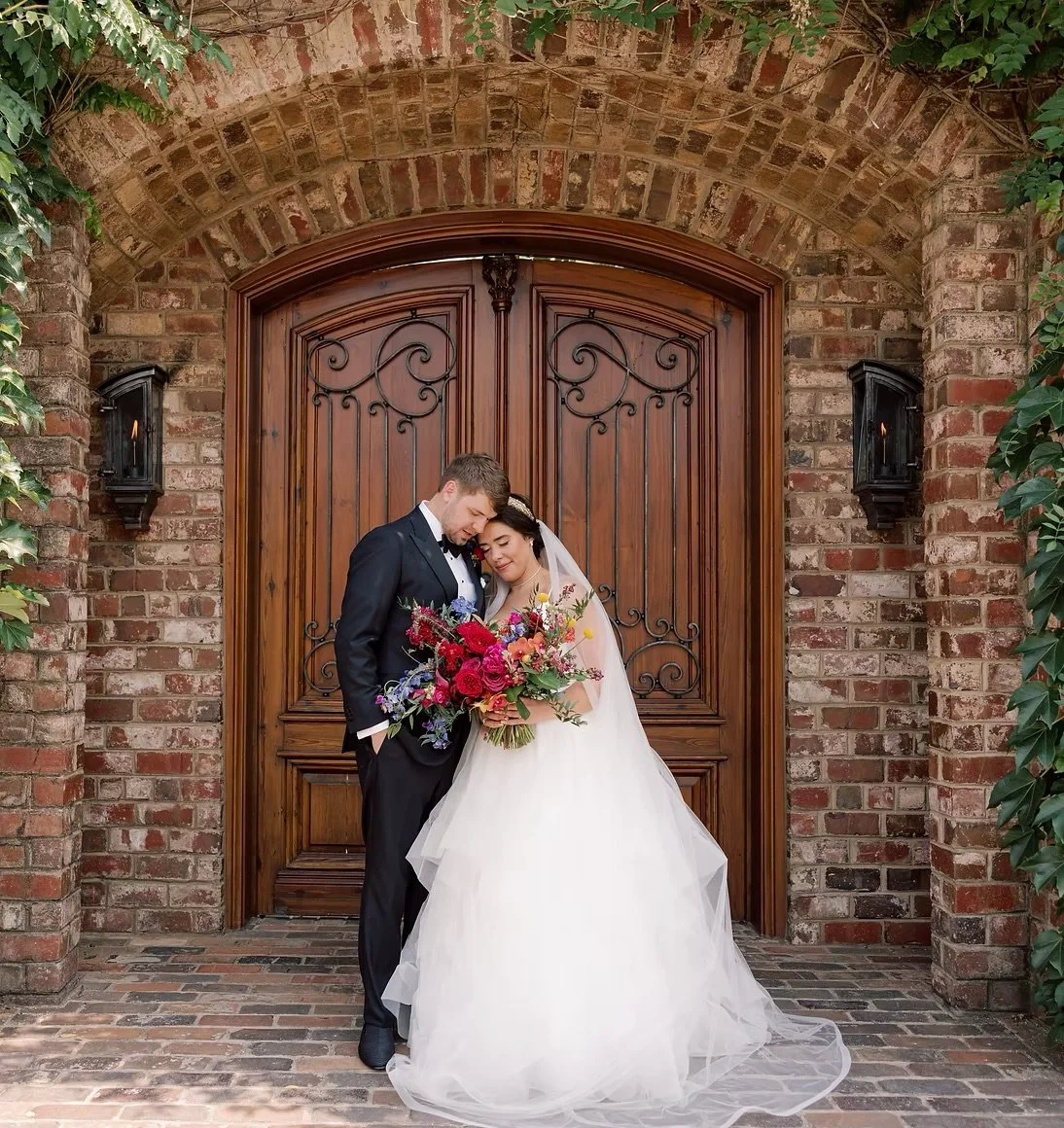 A bride and groom standing close together in front of a large wooden door, with brick walls and greenery around. The bride holds a vibrant bouquet of flowers and both have their heads touching in a tender moment.