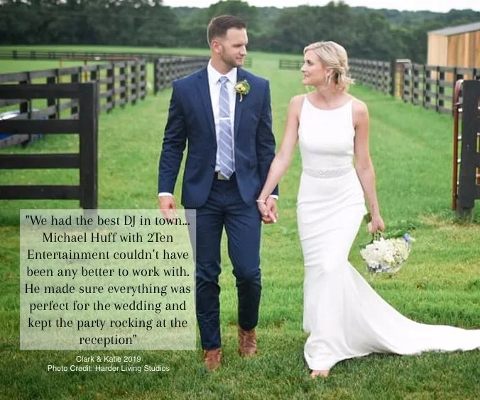 A bride and groom walking hand in hand outdoors at a wedding venue, with green grass and fencing in the background. The bride is wearing a white wedding dress and holding a bouquet of flowers, while the groom is dressed in a navy blue suit and tie.