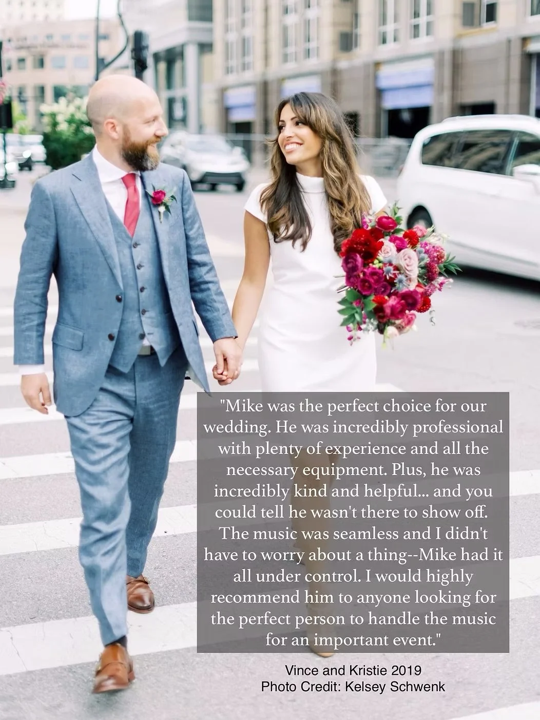 A bride and groom walking hand in hand on a city street; the bride is holding a bouquet of pink and red flowers, and the groom is wearing a blue suit with a red tie and boutonniere.