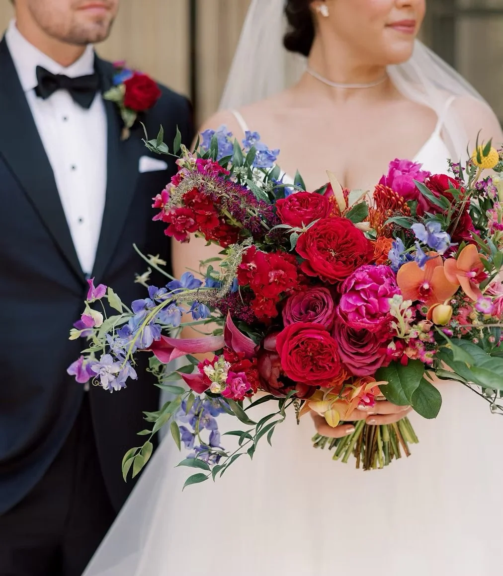 Bride holding a large, colorful bouquet of flowers with a groom in a tuxedo standing beside her.