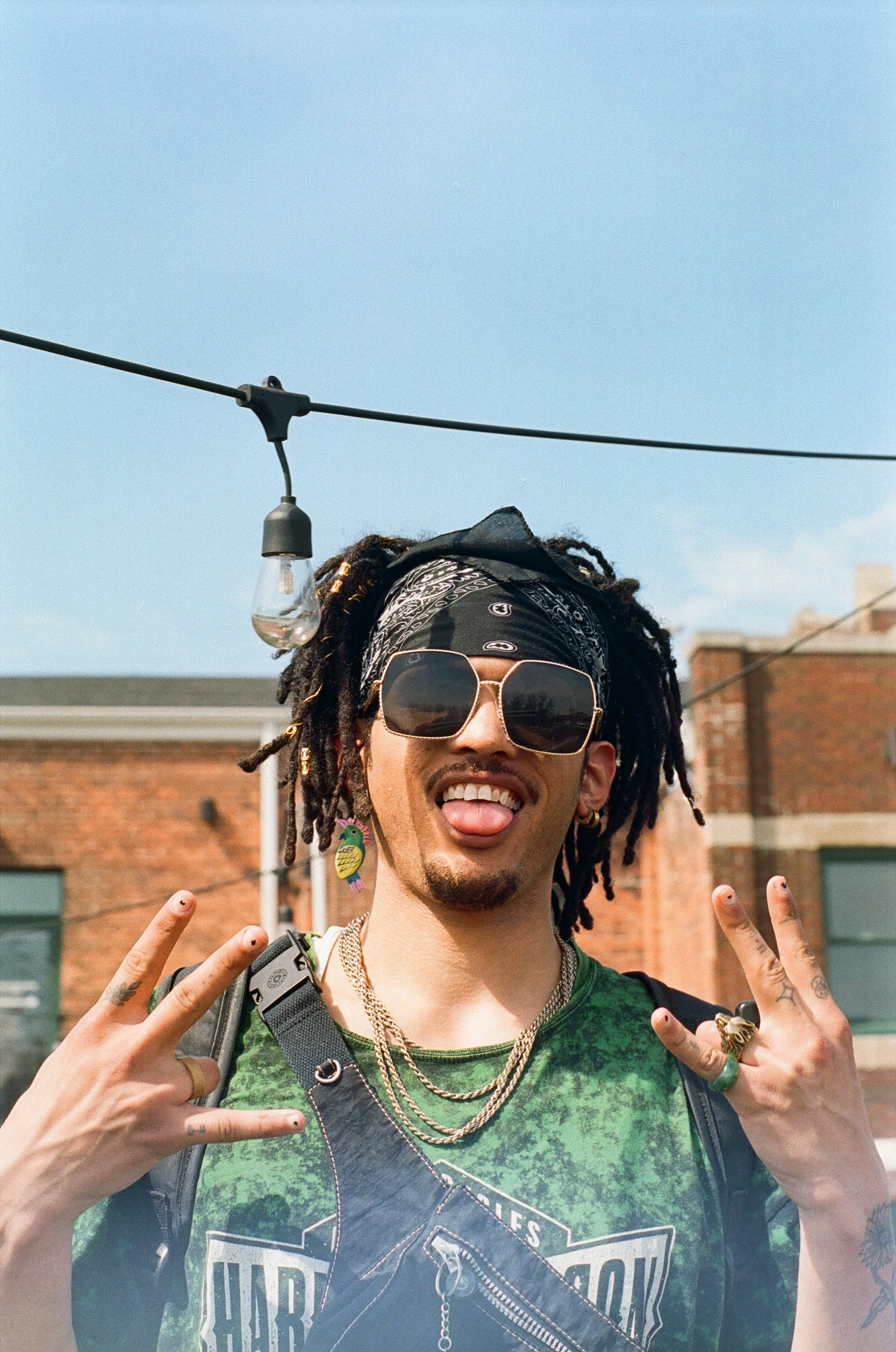 A person with dreadlocks wearing sunglasses, a bandana, and colorful earrings, making a peace sign with both hands outdoors under string lights on a sunny day.