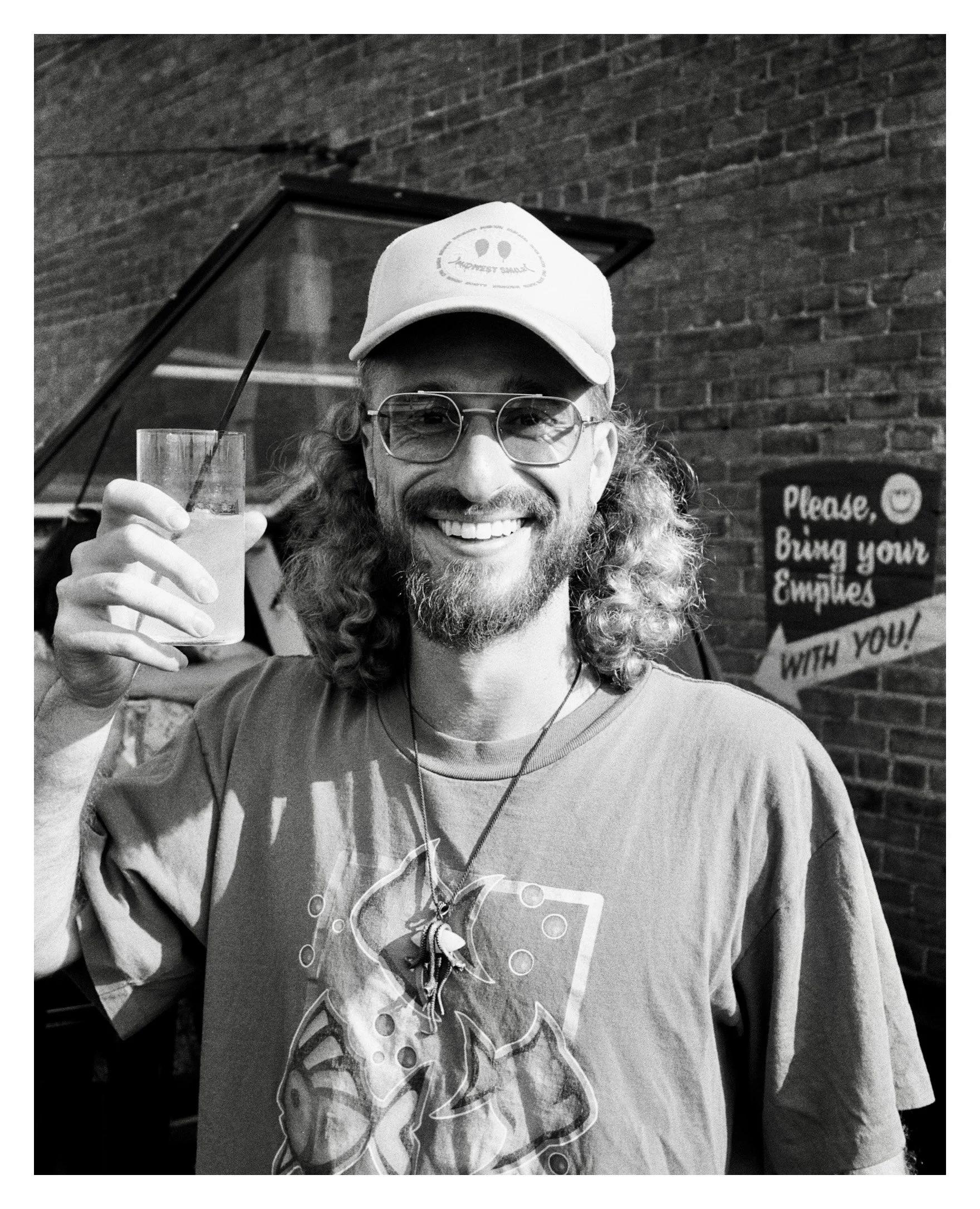 A man with shoulder-length curly hair, glasses, and a beard smiling while holding a drink with a straw in front of a brick wall and a sign that reads, 'Please, bring your empires with you!'