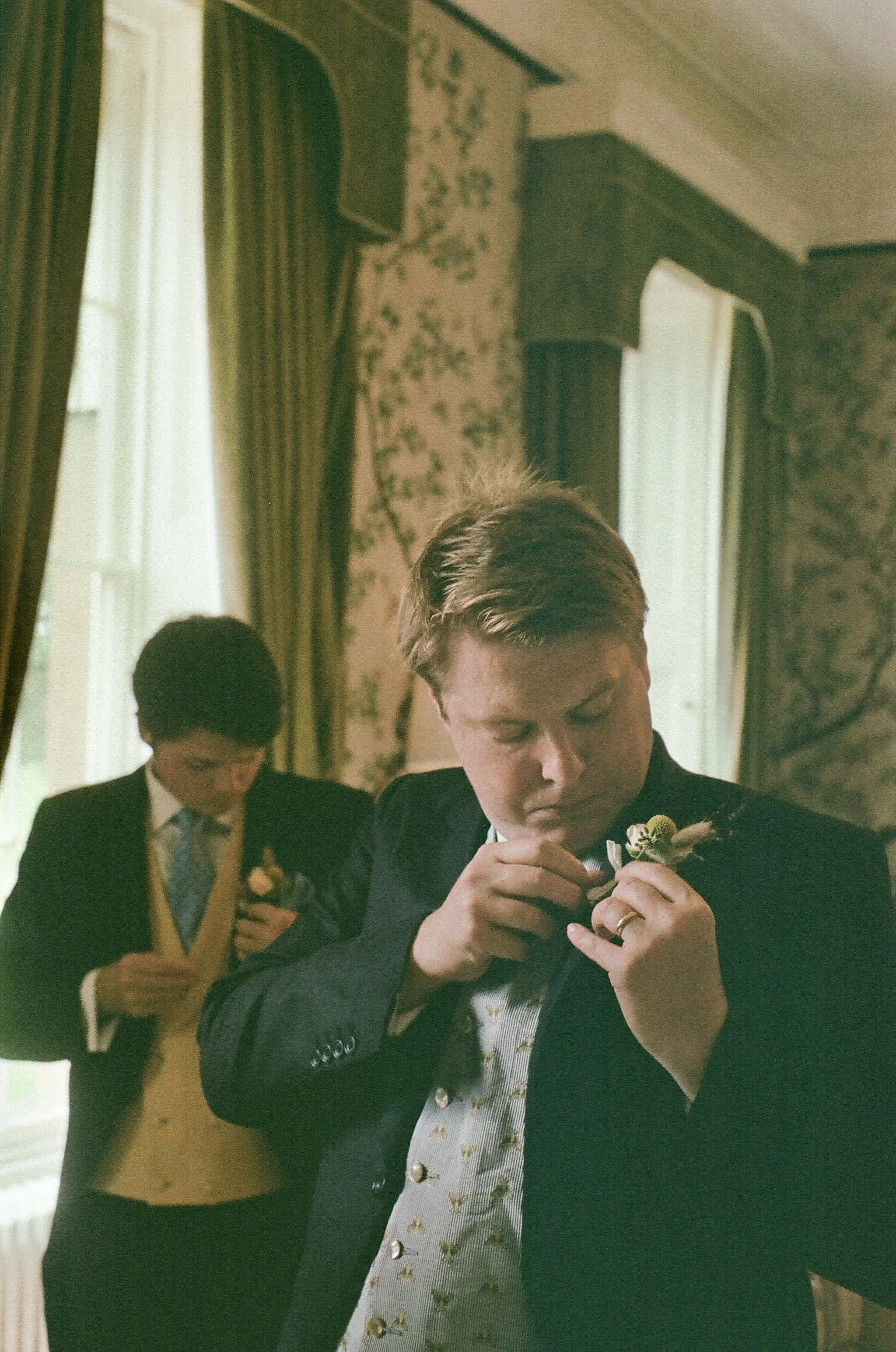 Two men in formal suits, one adjusting a boutonniere and the other preparing a boutonniere in a decorated room with curtains and floral wallpaper.