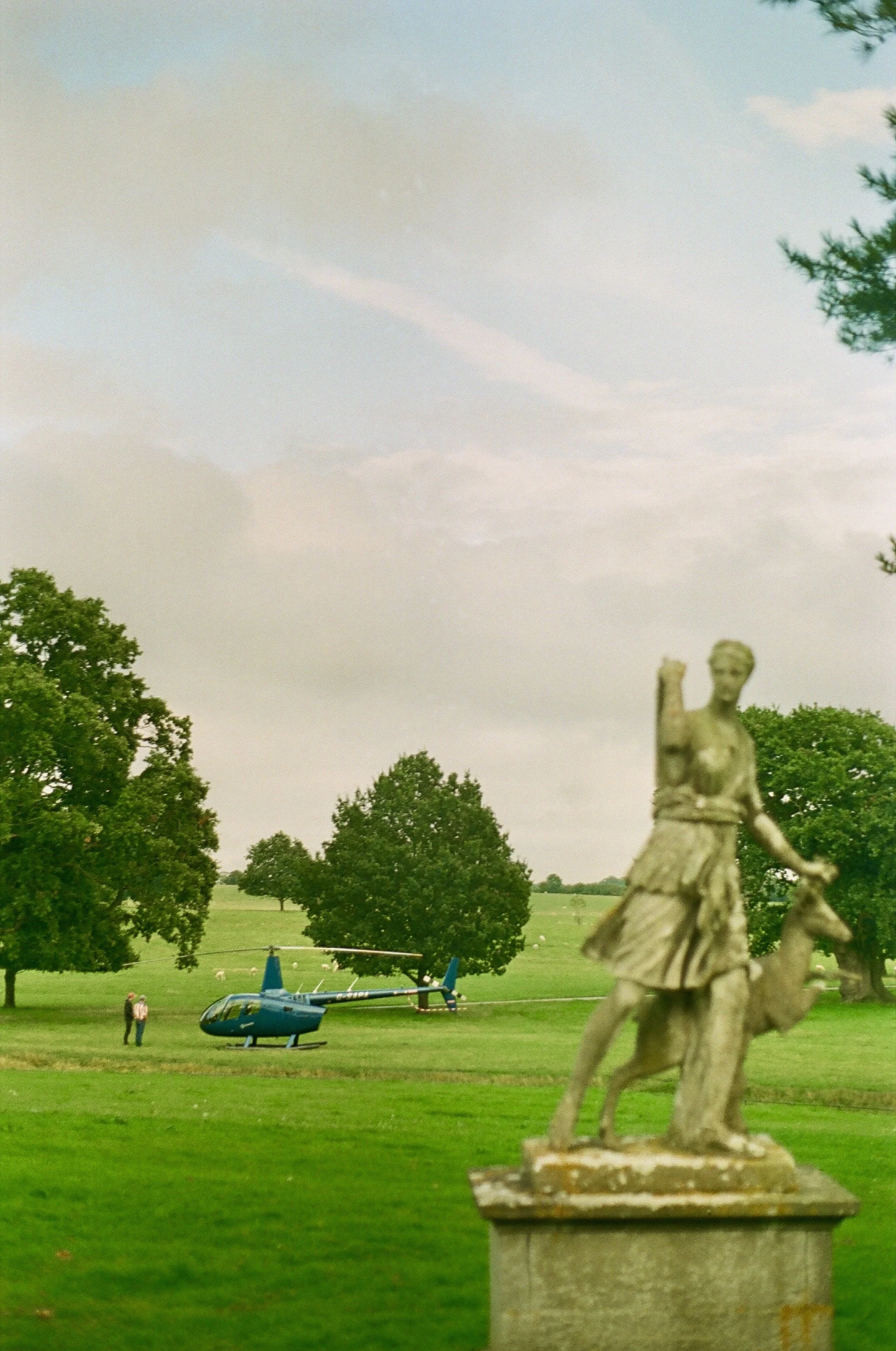 A historic stone sculpture of a woman with a dog in the foreground, with a helicopter, trees, and an open field in the background under a partly cloudy sky.