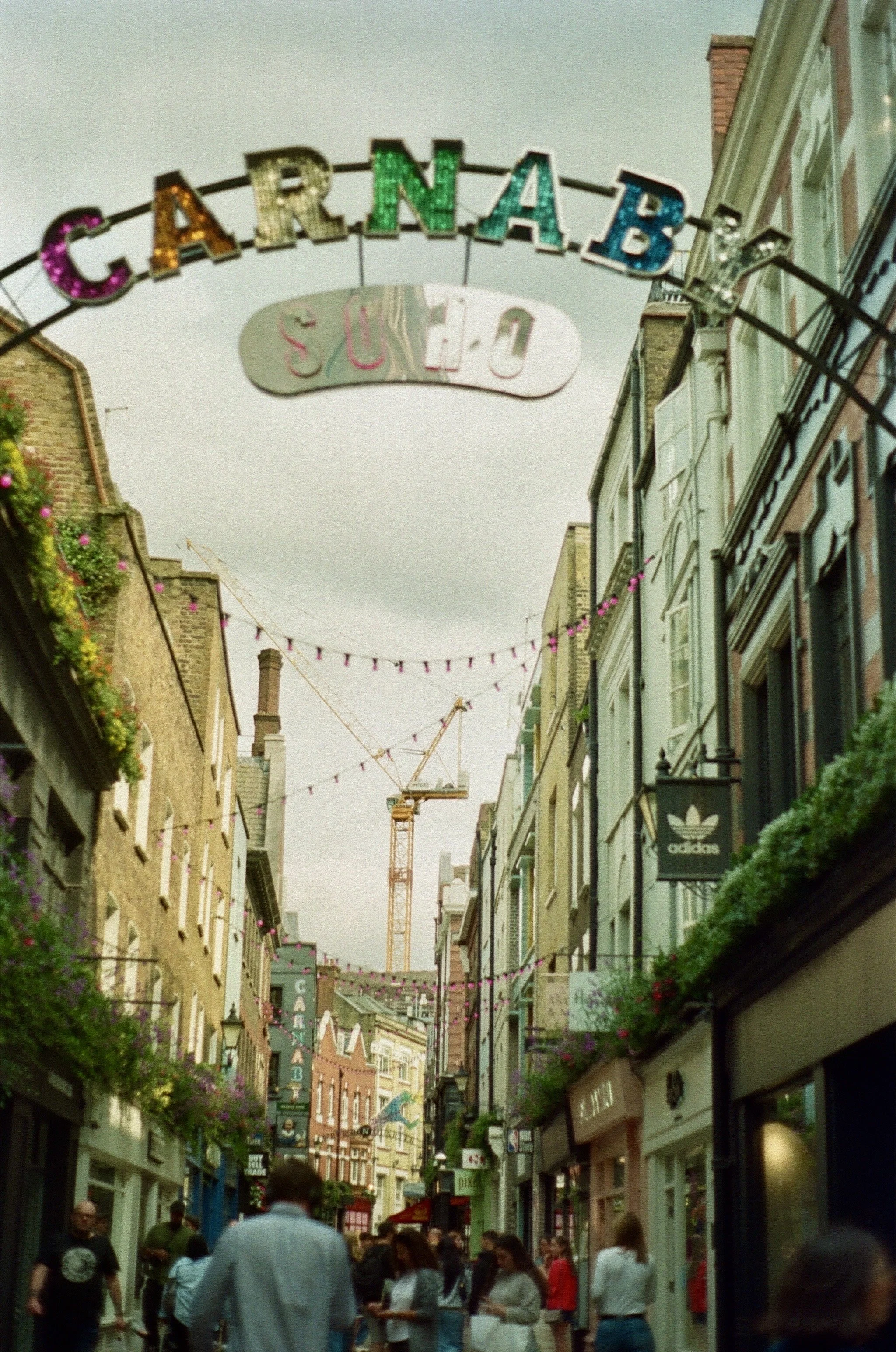 A busy city street with colorful signs, hanging pink string lights, and a construction crane in the background, leading to a carnival sign.