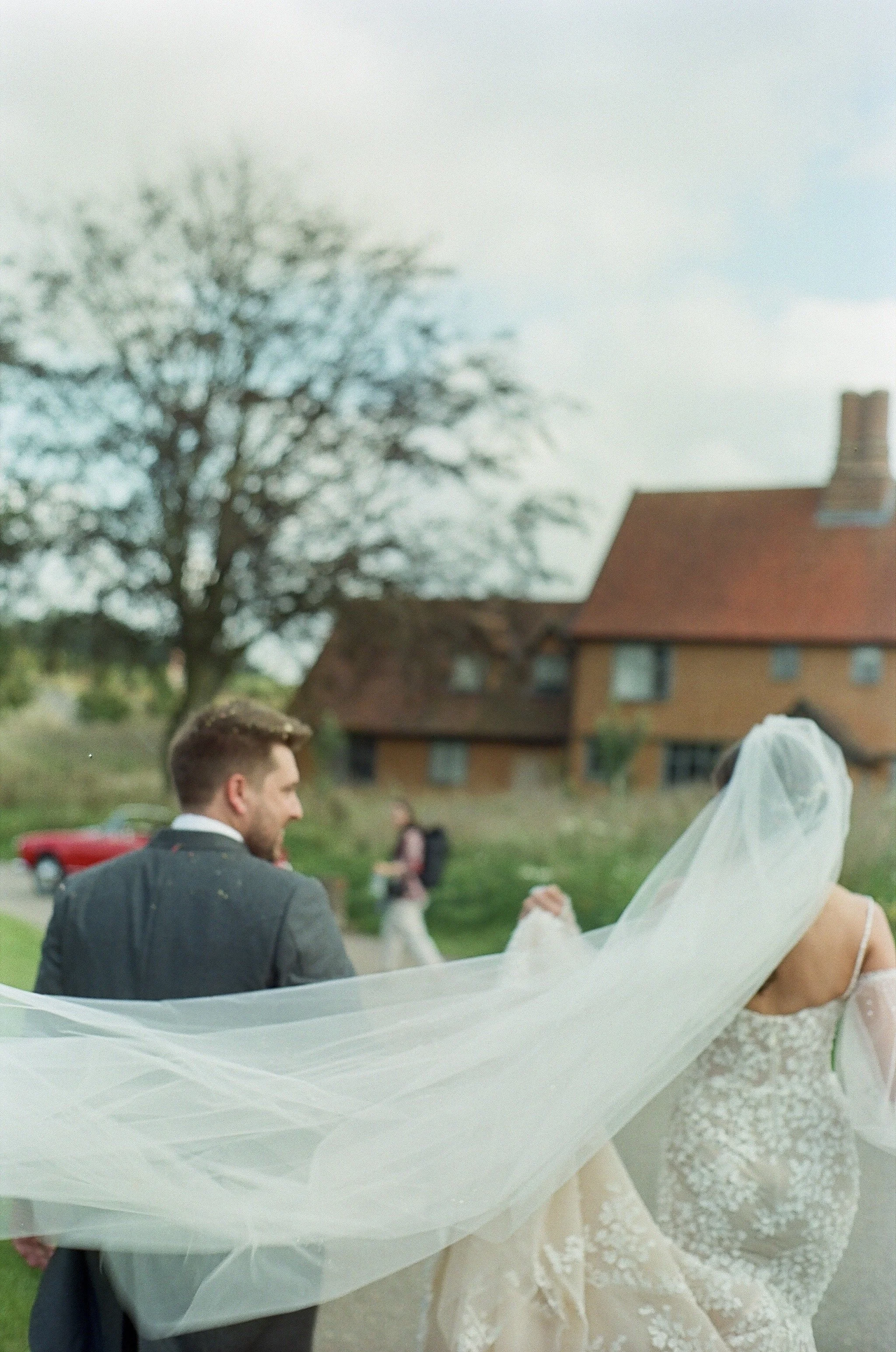 A bride and groom holding hands and dancing outdoors in front of a large house with a red roof, with a person in the background.