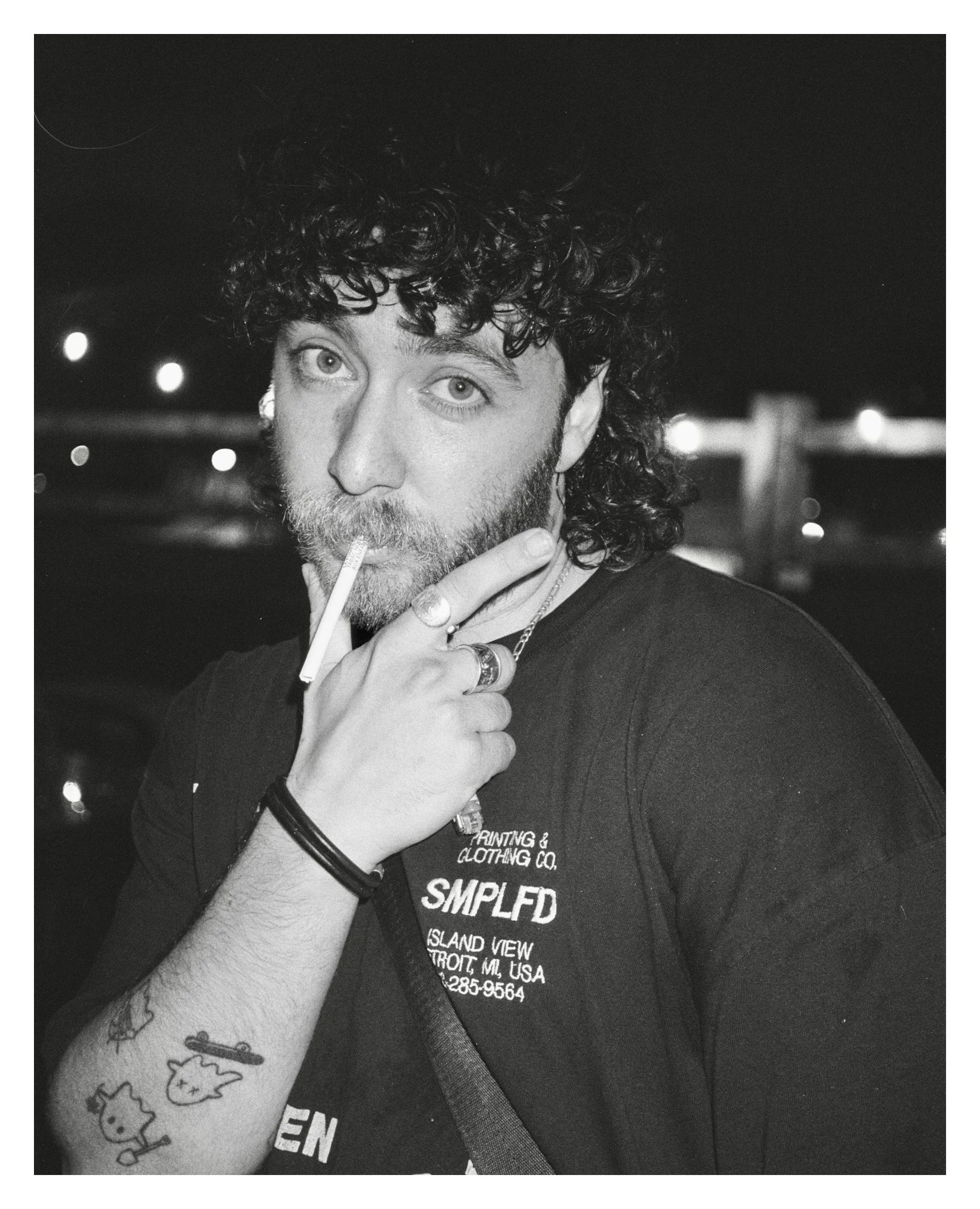 A man with curly hair and a beard smoking a cigarette at night, wearing a black T-shirt with white text, jewelry, and tattoos on his arm.