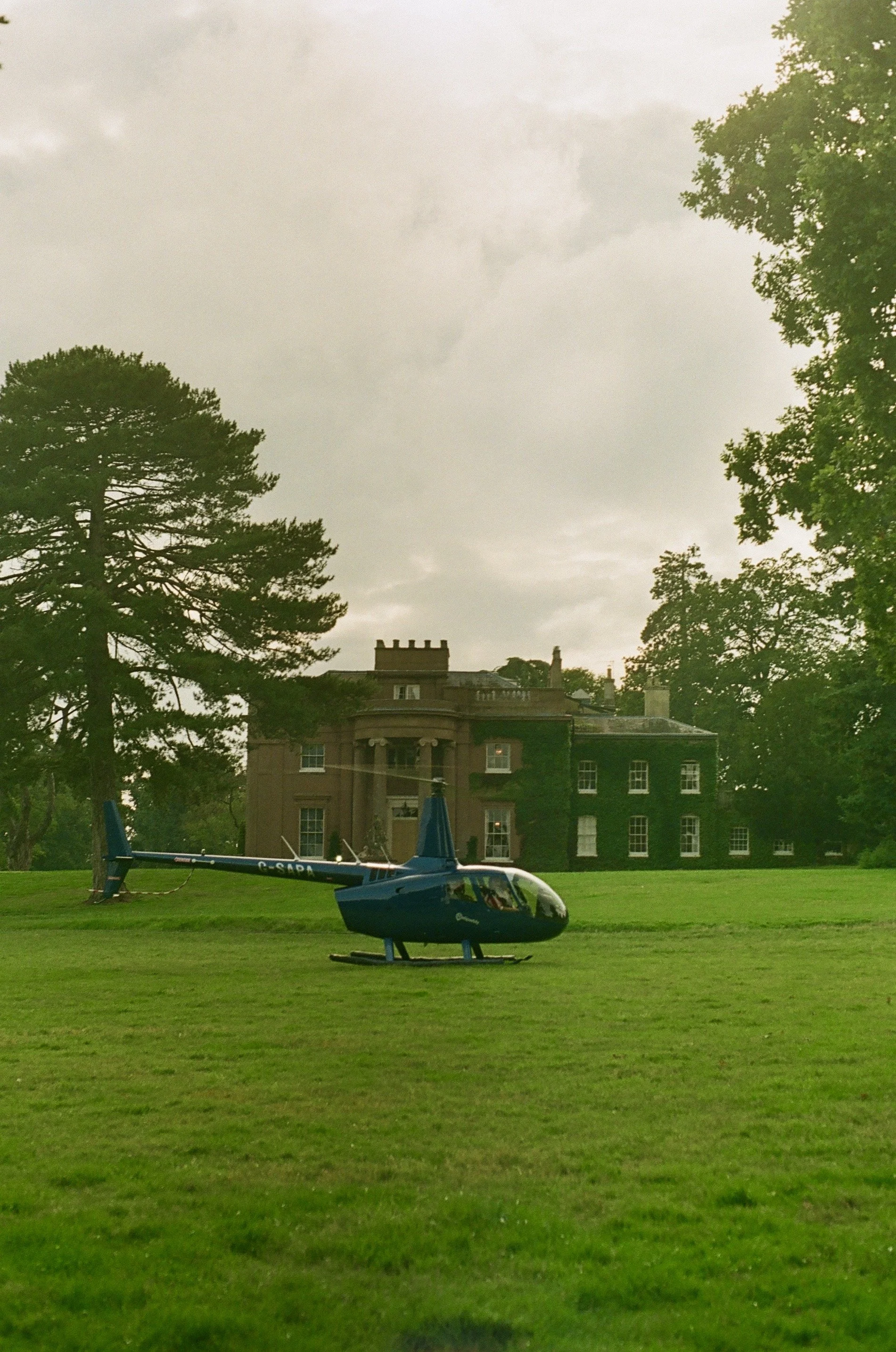 A blue helicopter on a grassy lawn in front of a large, historic mansion surrounded by tall trees and a cloudy sky.
