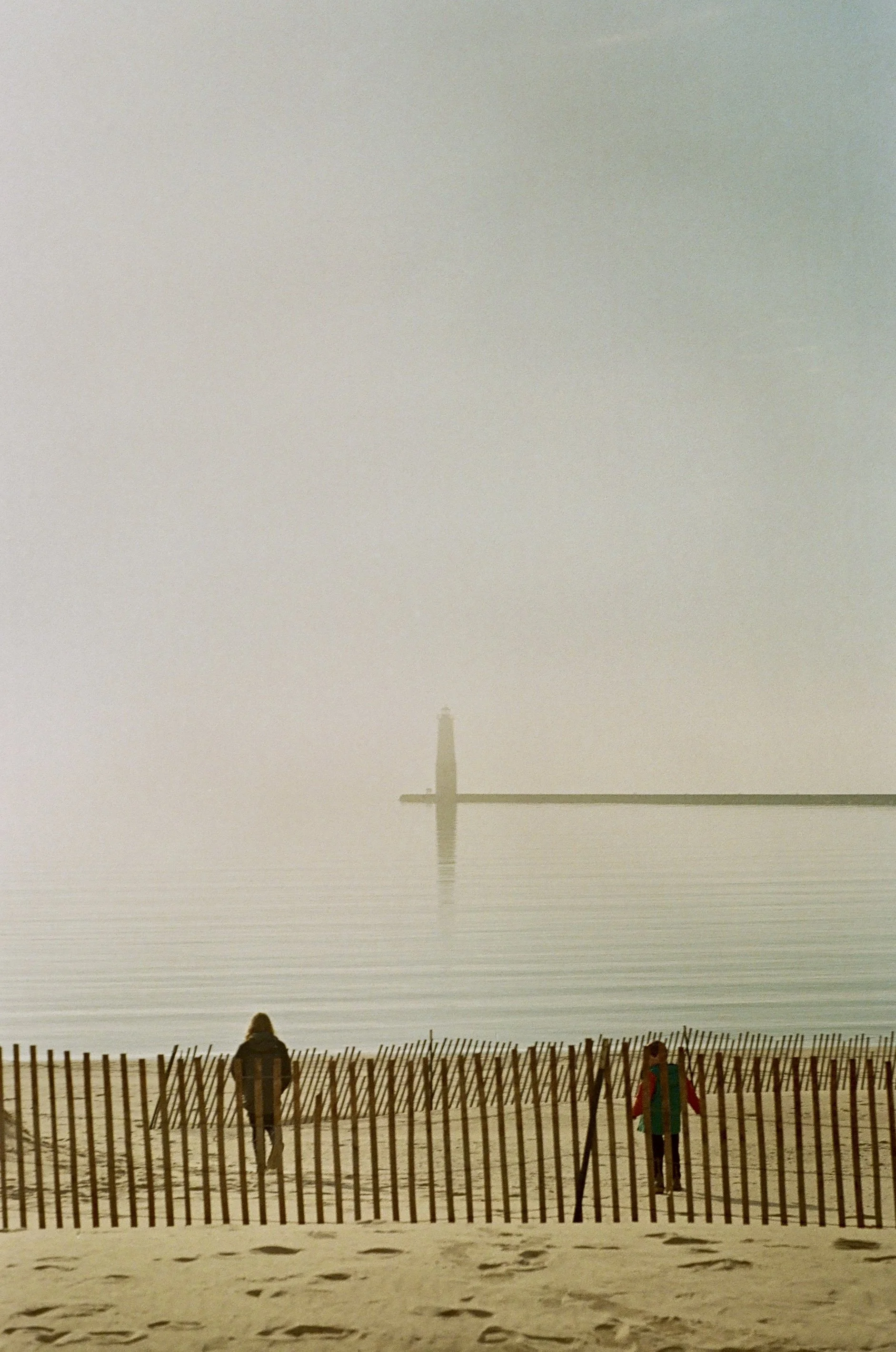 A person walking on a sandy beach near a wooden fence, with foggy weather and a lighthouse in the distance across calm water.