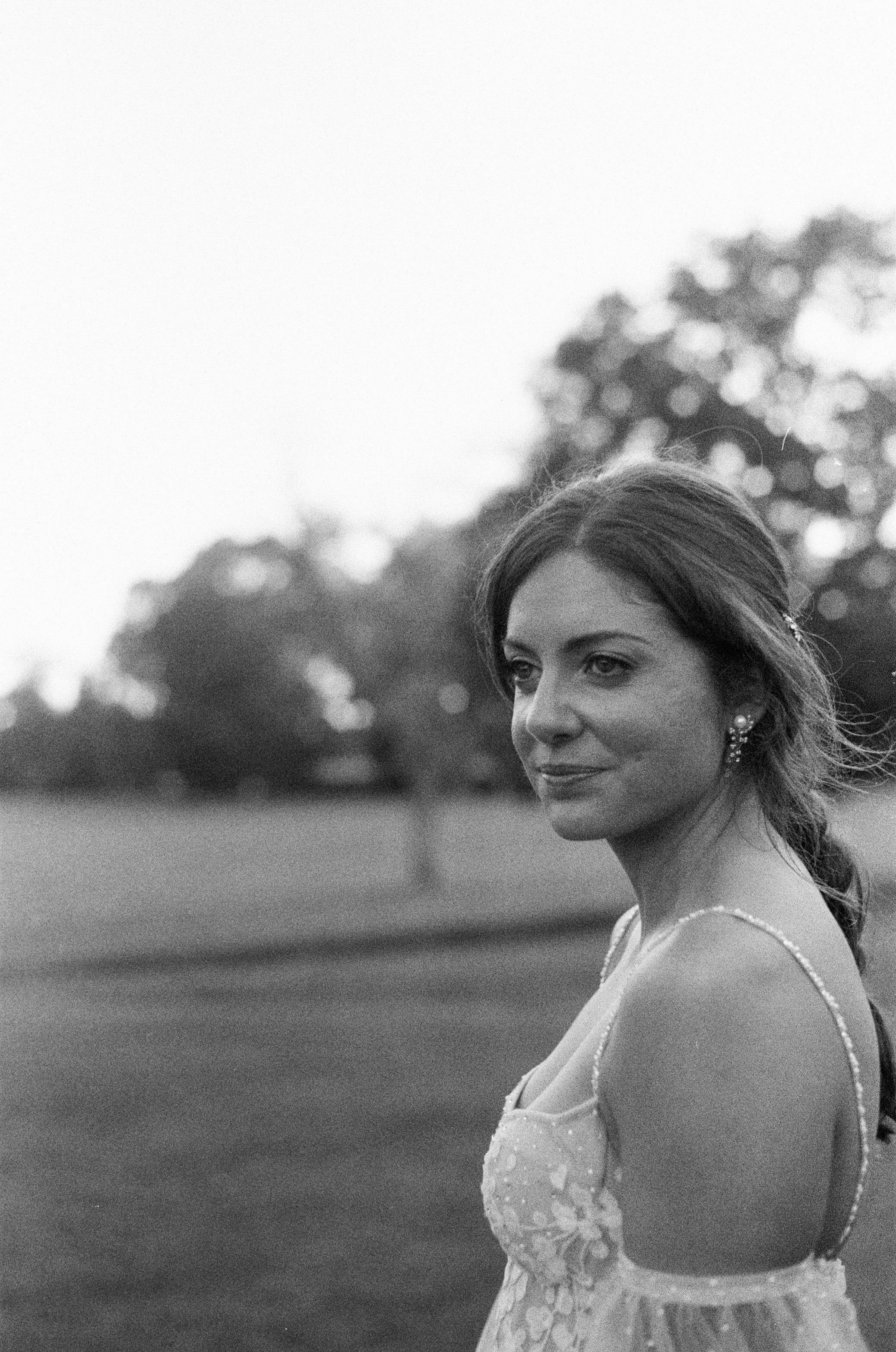 Black and white photo of a woman in a dress outdoors, looking to the side with a slight smile, against a blurred background of trees.