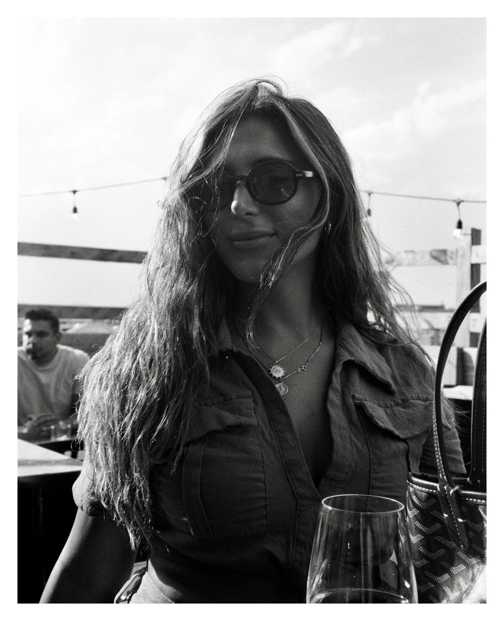 A woman with wavy hair wearing sunglasses and a denim jacket, sitting outdoors at a table with a glass of wine in front of her, with string lights and a man in the background.