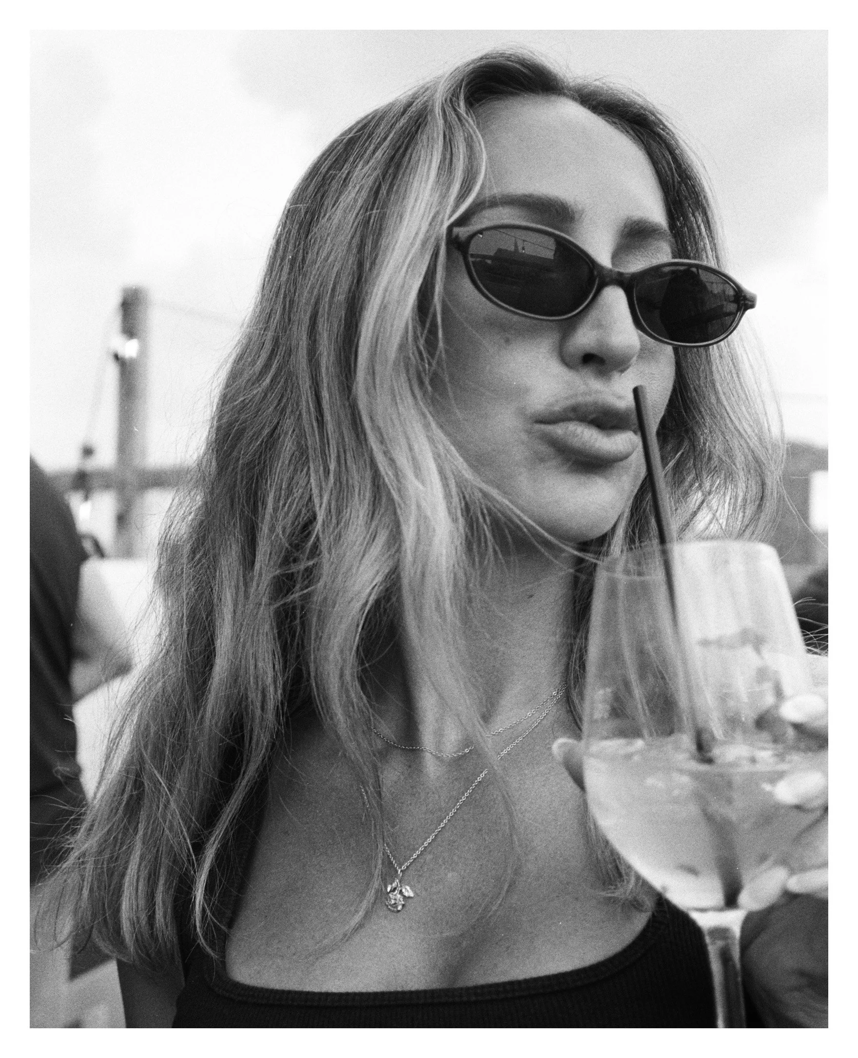 A woman with long, wavy hair wearing sunglasses sips a drink through a straw at an outdoor setting.