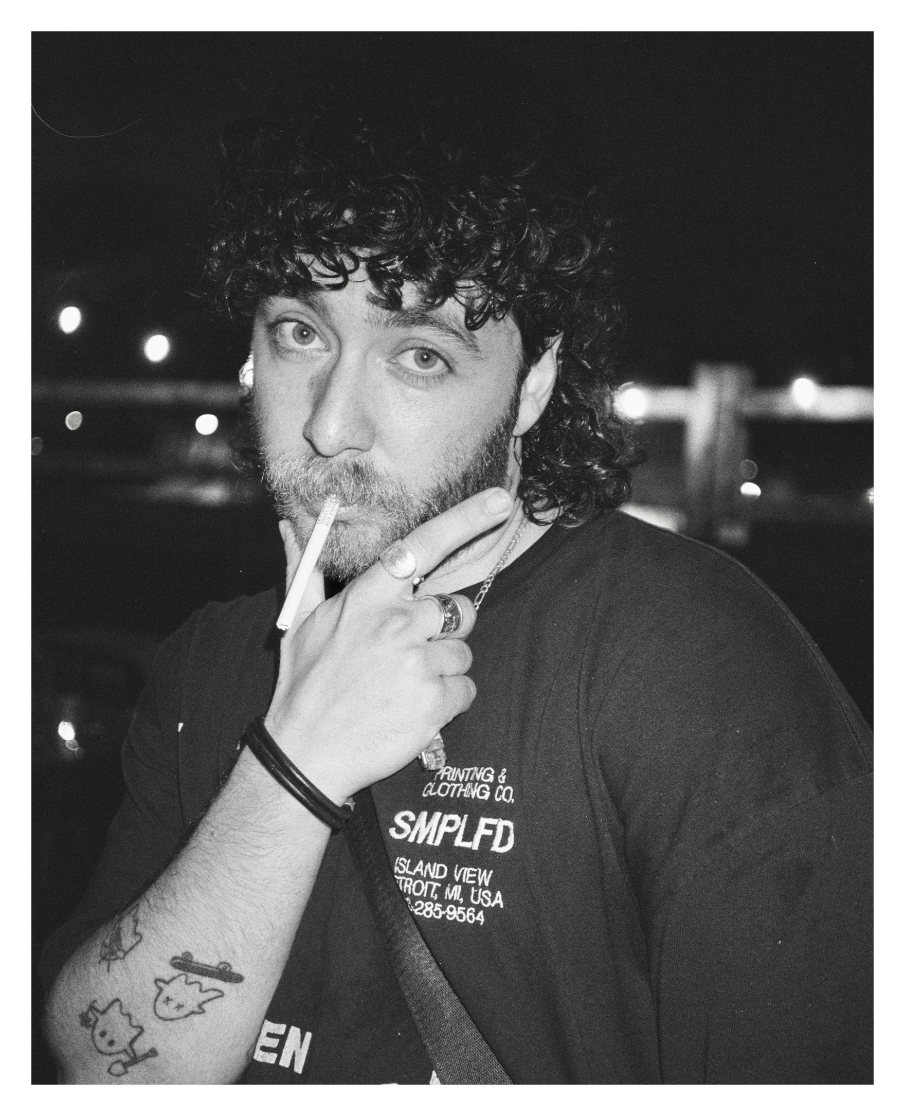 A young man with curly hair and a beard holding a cigarette to his lips with his fingers, wearing a black T-shirt, rings, and a bracelet, standing outdoors at night with blurred lights in the background.