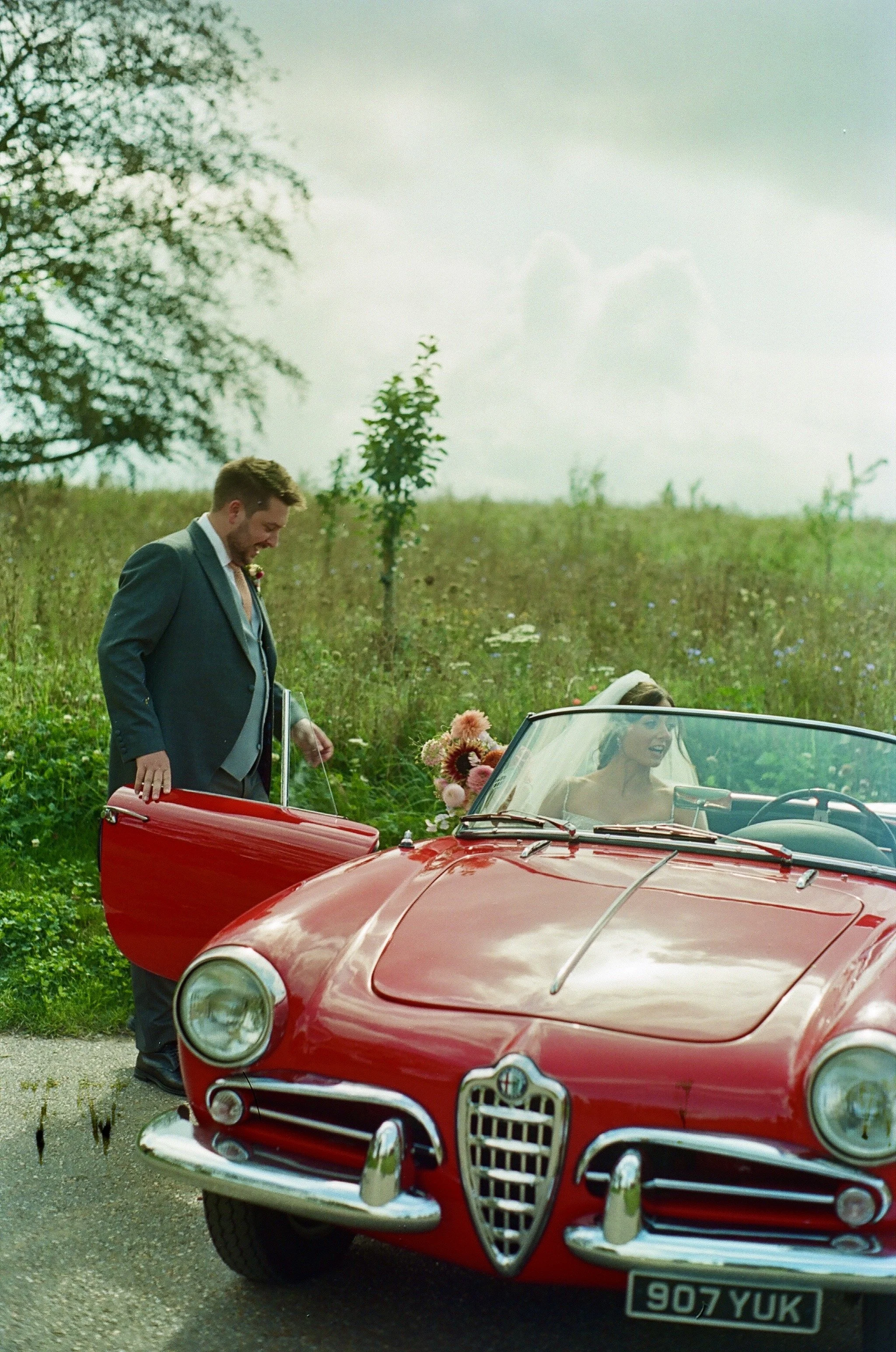 A bride and groom with a vintage red Alfa Romeo convertible car outdoors on a grassy field, the groom opening the car door while the bride sits inside, holding a bouquet of flowers, with trees and a cloudy sky in the background.