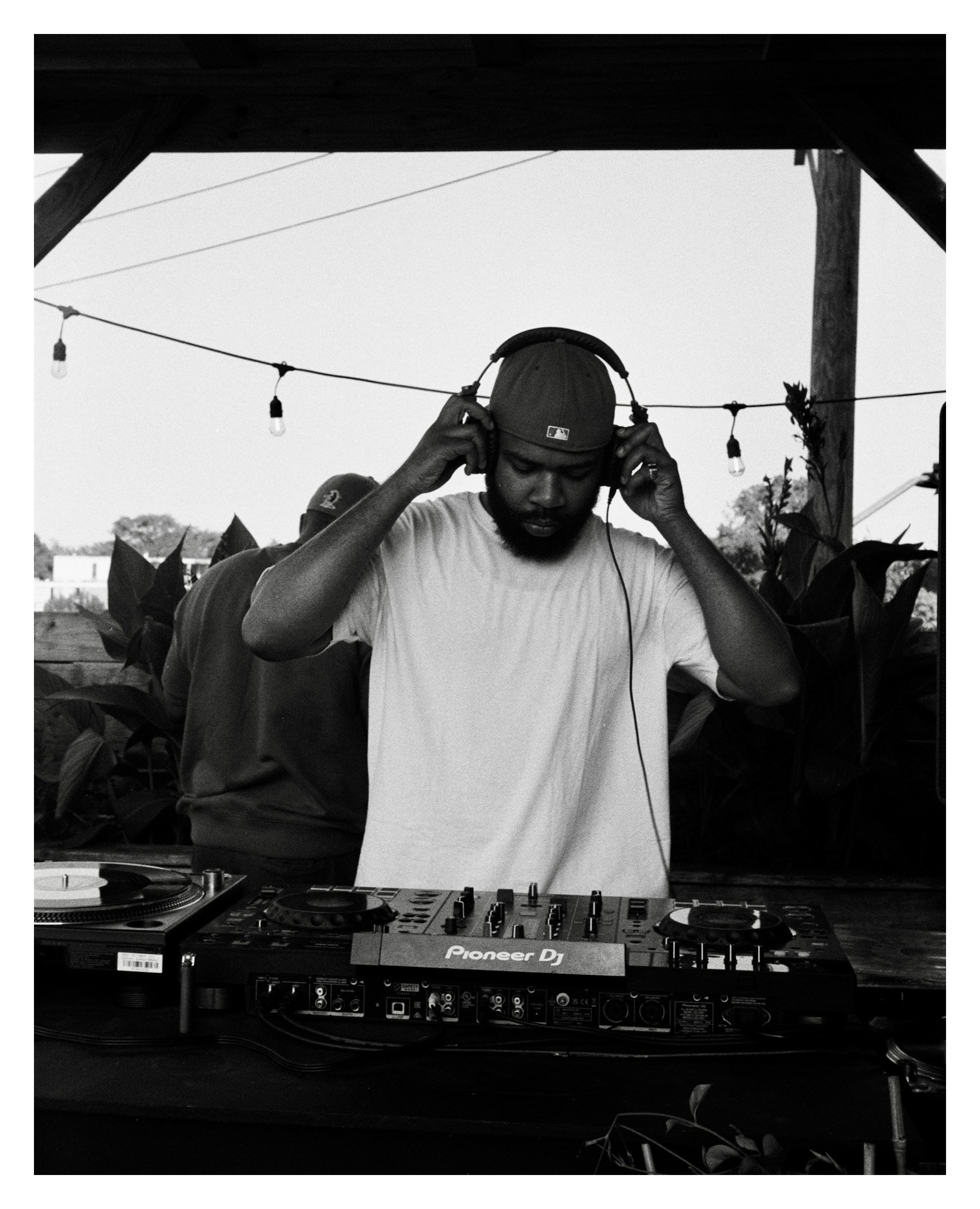 A man with a beard and cap wearing headphones, standing behind DJ equipment outdoors.