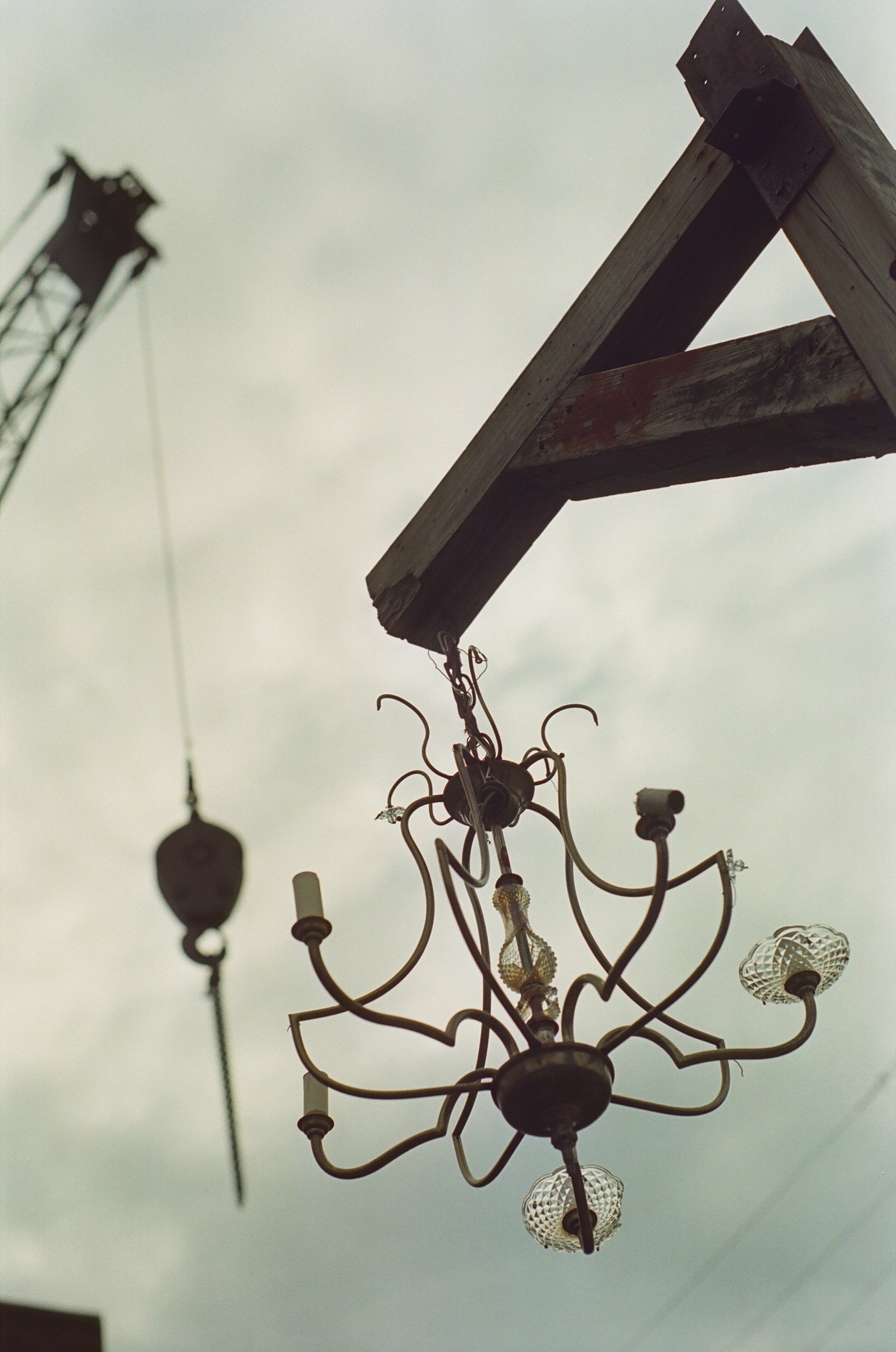 An old chandelier hanging from a wooden beam against a cloudy sky, with a blurred crane visible in the background.