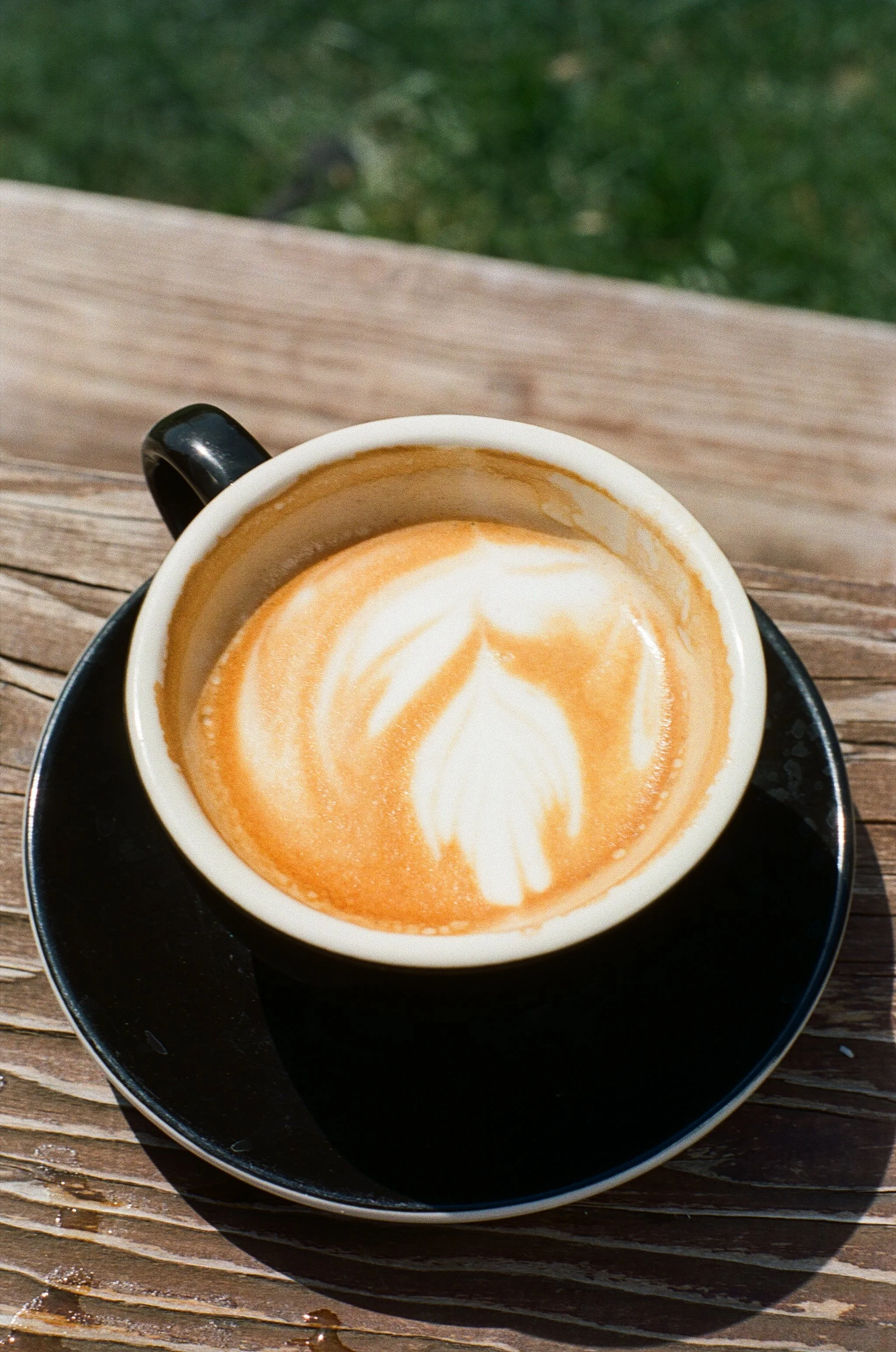 A cup of latte with leaf-shaped foam art on top, sitting on a black saucer on a wooden surface outdoors.