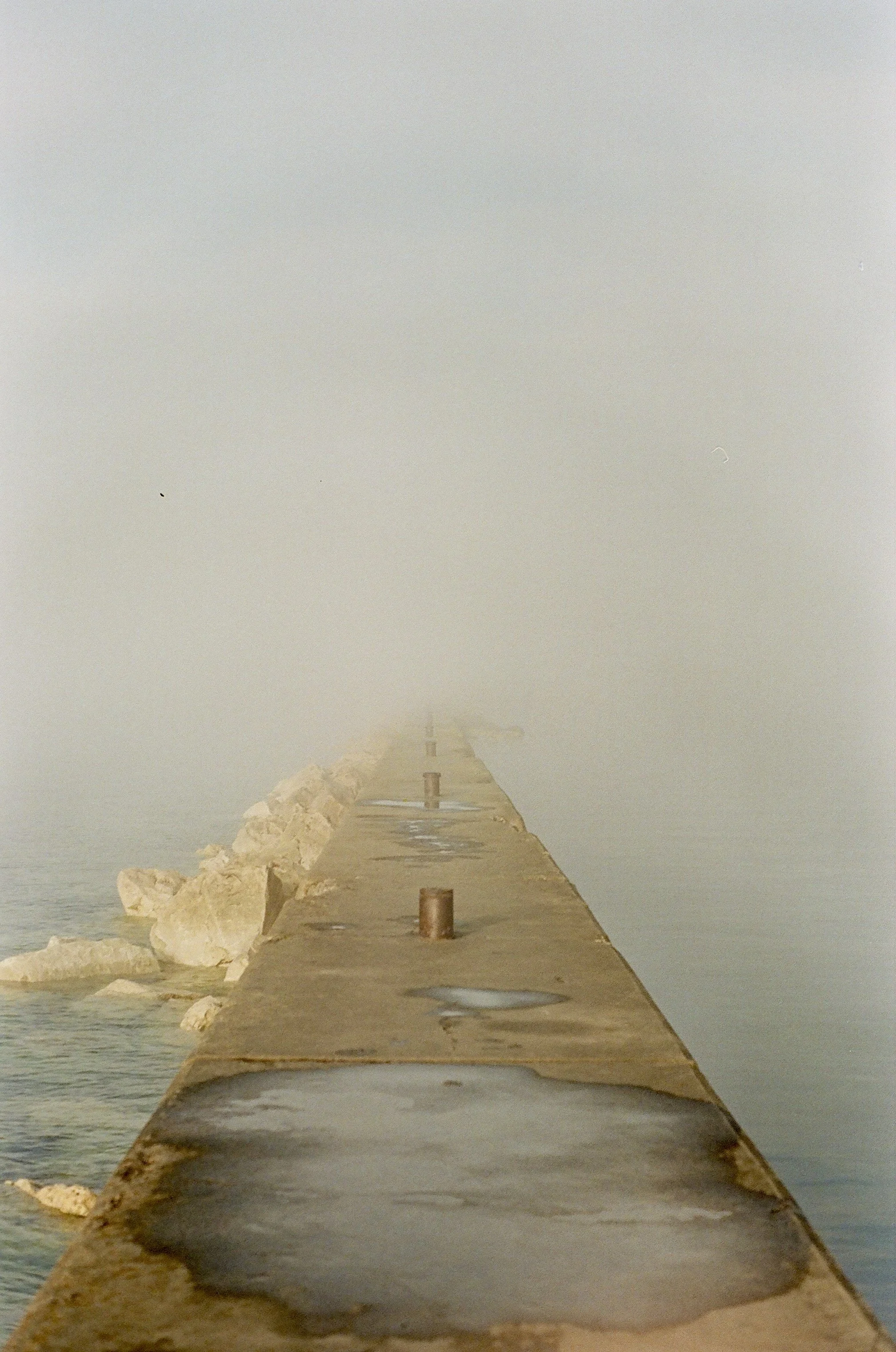 A foggy pier extending into a body of water with rocks on the left side and rusty bollards along the pier.