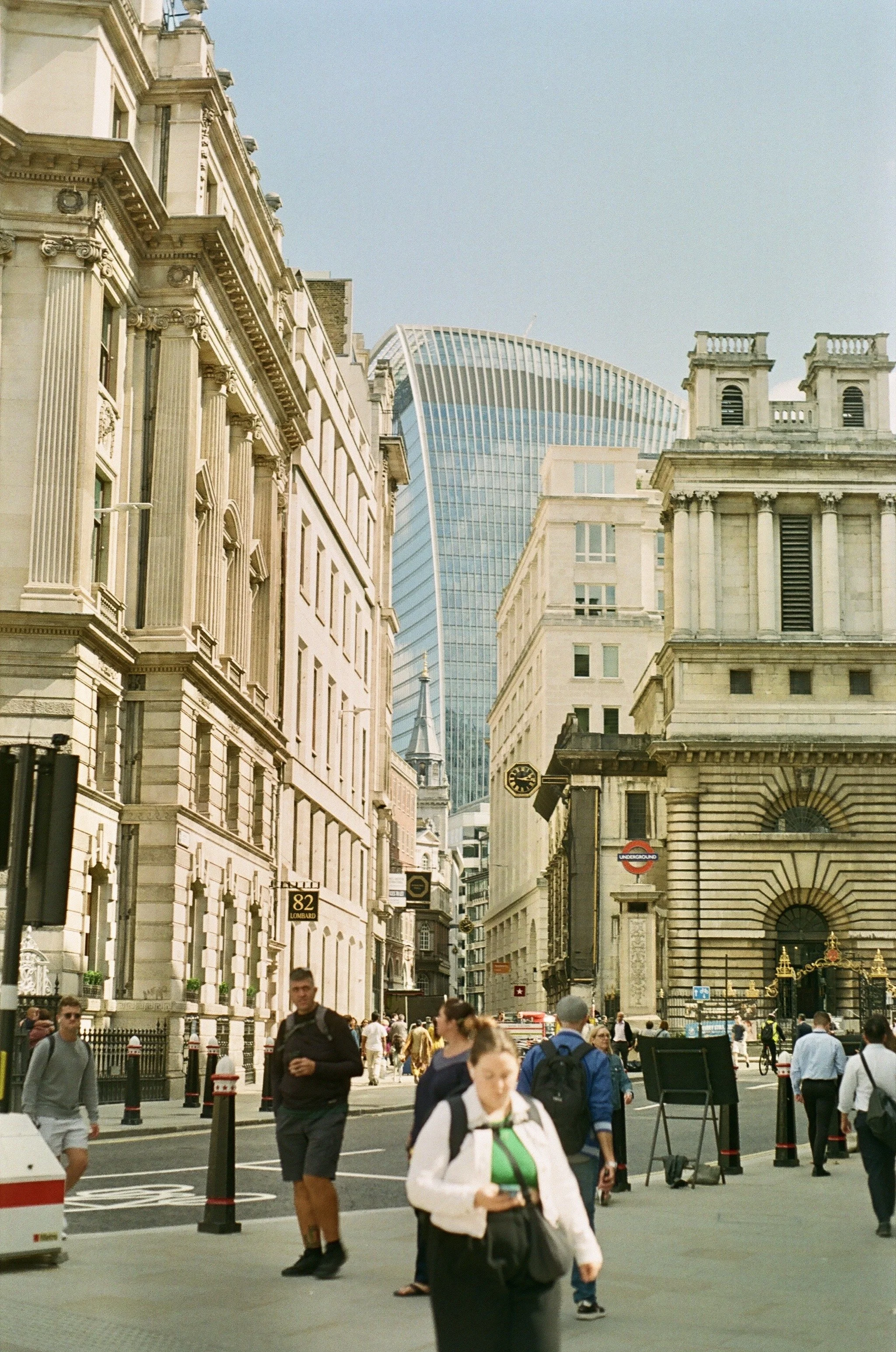 Street scene in London with diverse pedestrians, historic and modern buildings, and a partly cloudy sky.