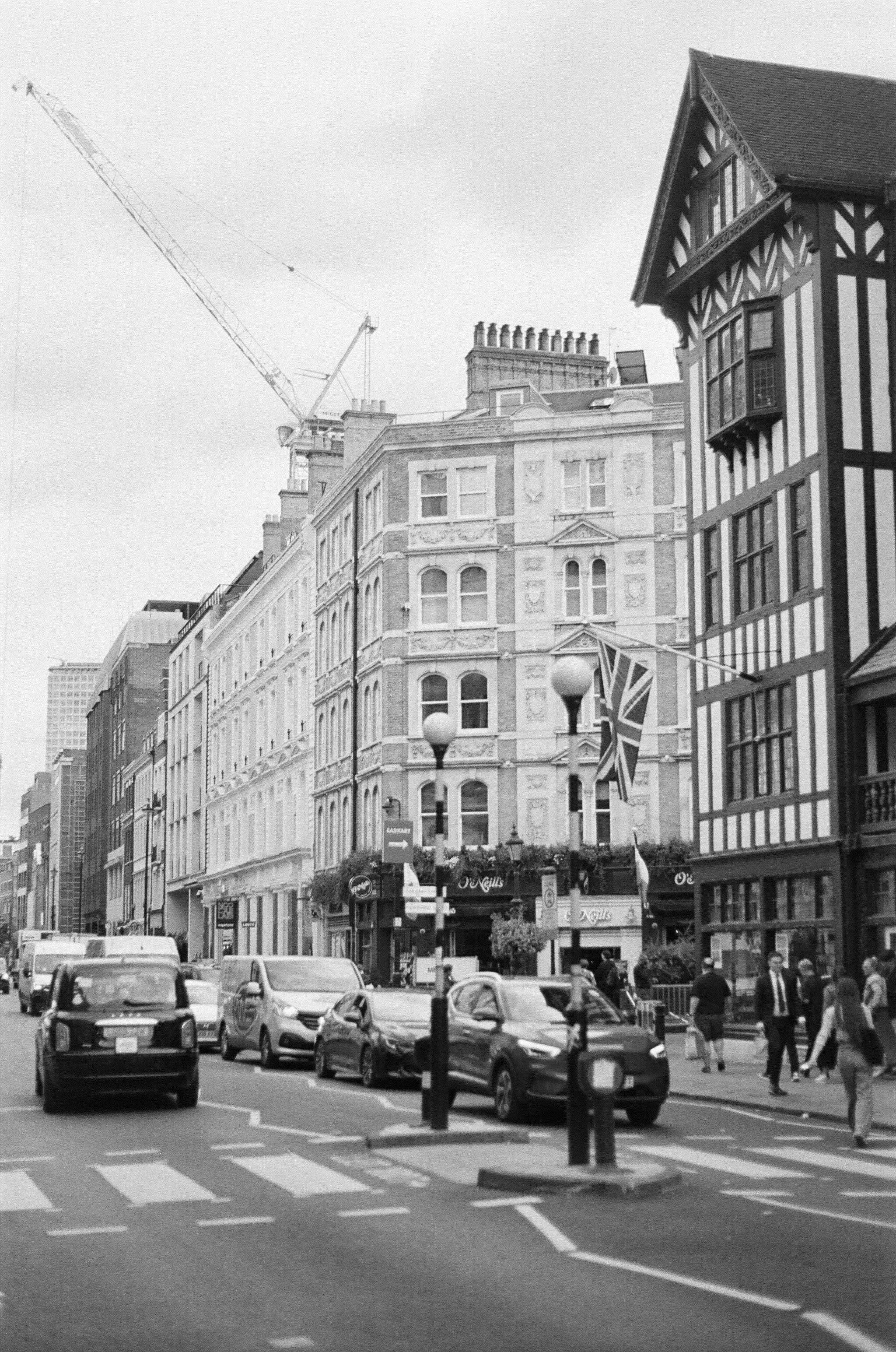 Black and white photo of city street with historic and modern buildings, parked cars, pedestrians walking, and Union Jack flags hanging.