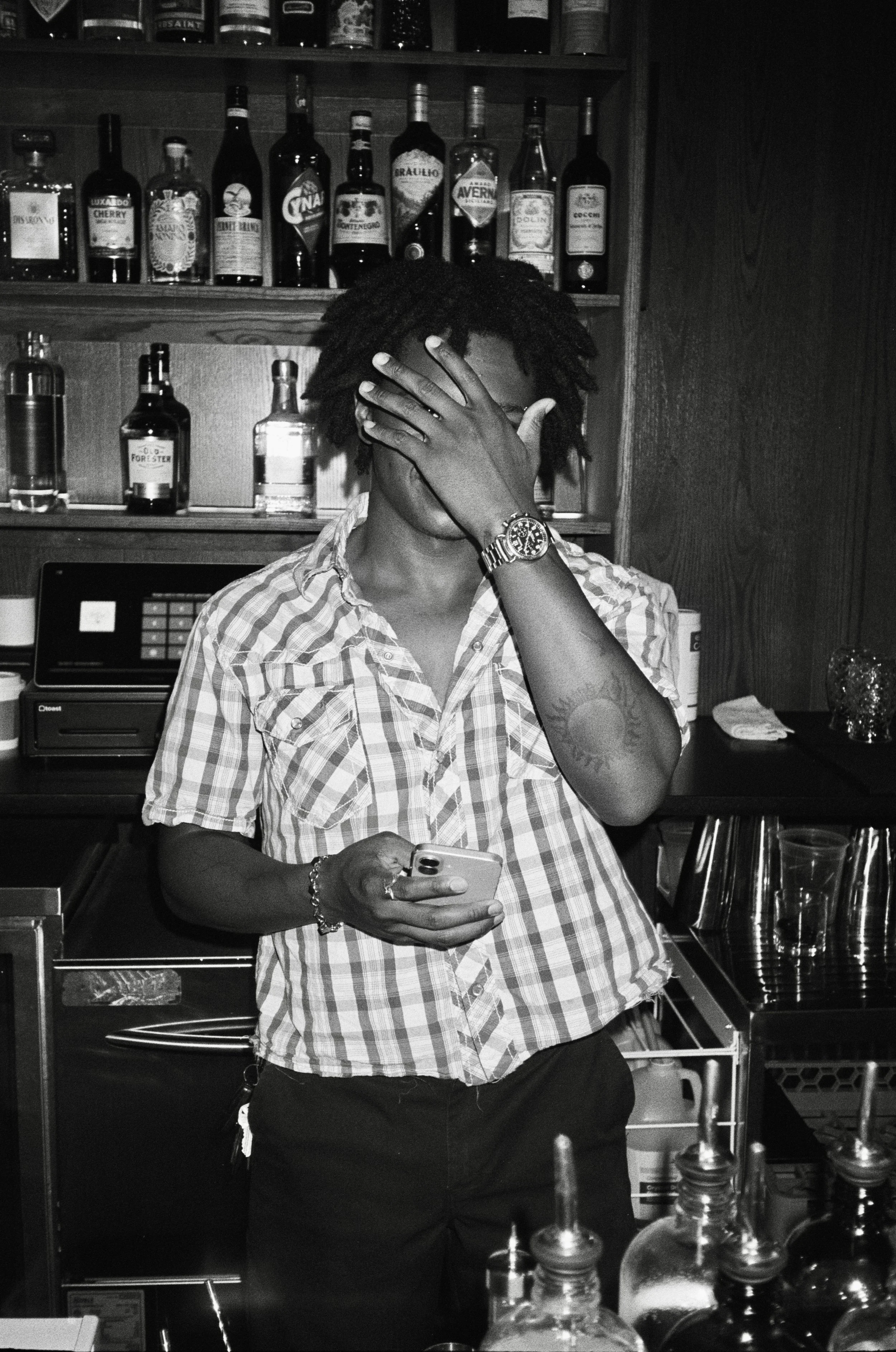 A person with dreadlocks covering their face, wearing a plaid shirt and a watch, standing behind a bar with liquor bottles on shelves in the background, holding a phone.