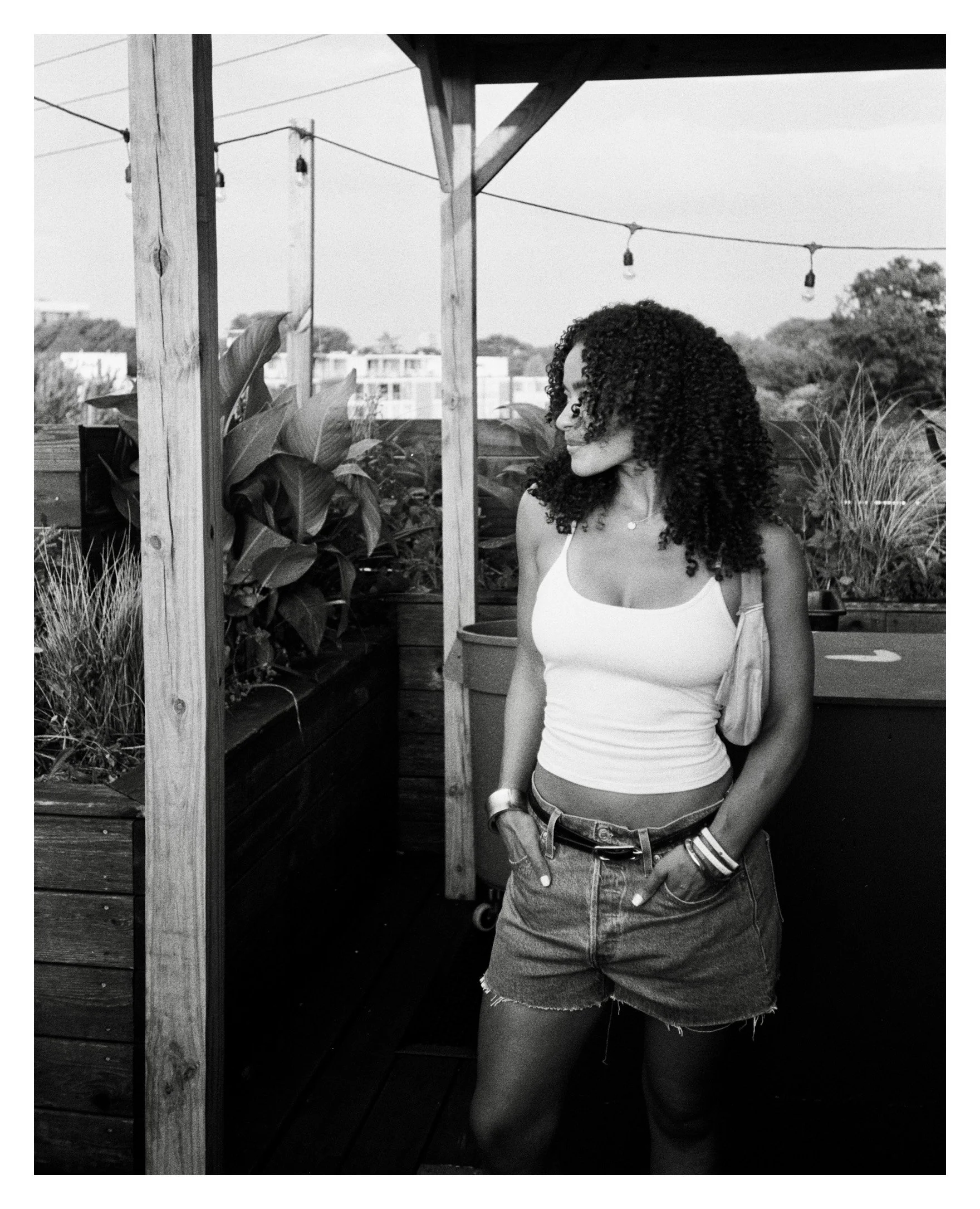 A woman with curly hair wearing a tank top and denim shorts standing outdoors on a wooden deck, surrounded by plants and string lights.