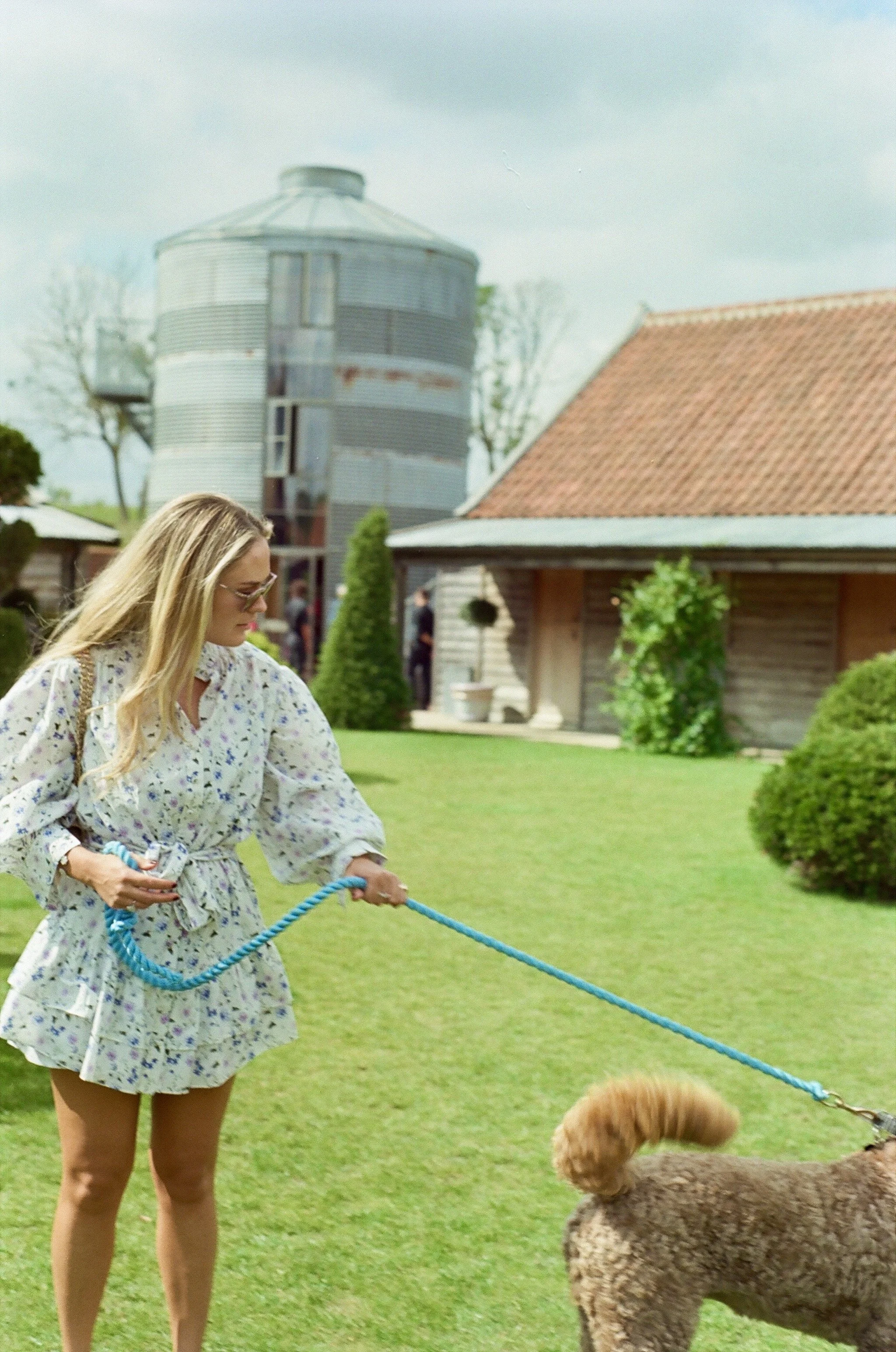 A woman with long blonde hair wearing sunglasses, a floral dress, and holding a blue leash, walking a curly-haired brown dog on a grassy lawn near a wooden building with a tiled roof, and a large metal grain silo in the background.