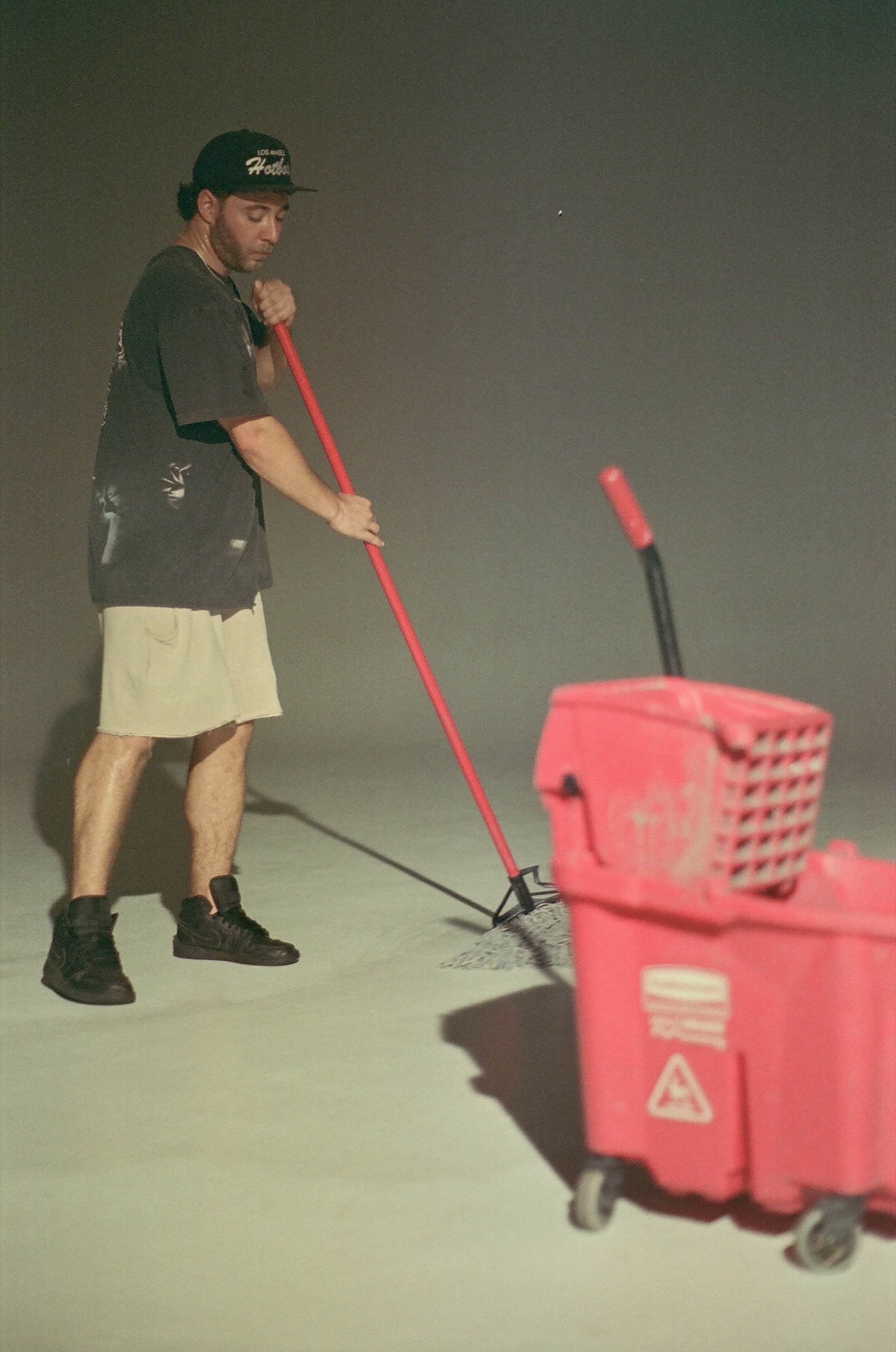 Young man mops a floor in a plain, empty room with dark background, using a red mop and a red cleaning cart.