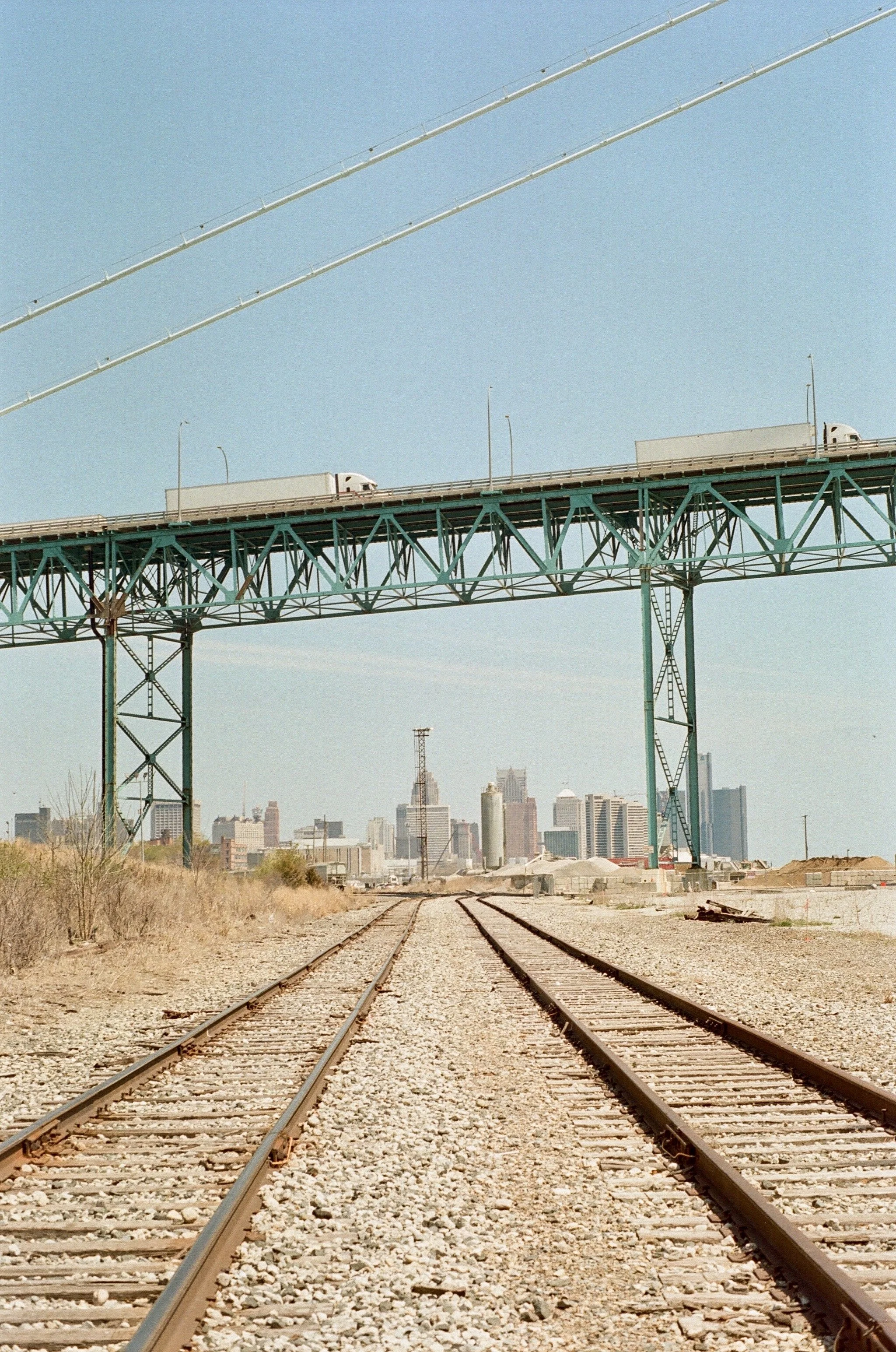Train tracks leading toward a city skyline with a bridge overhead and two trucks on the bridge.
