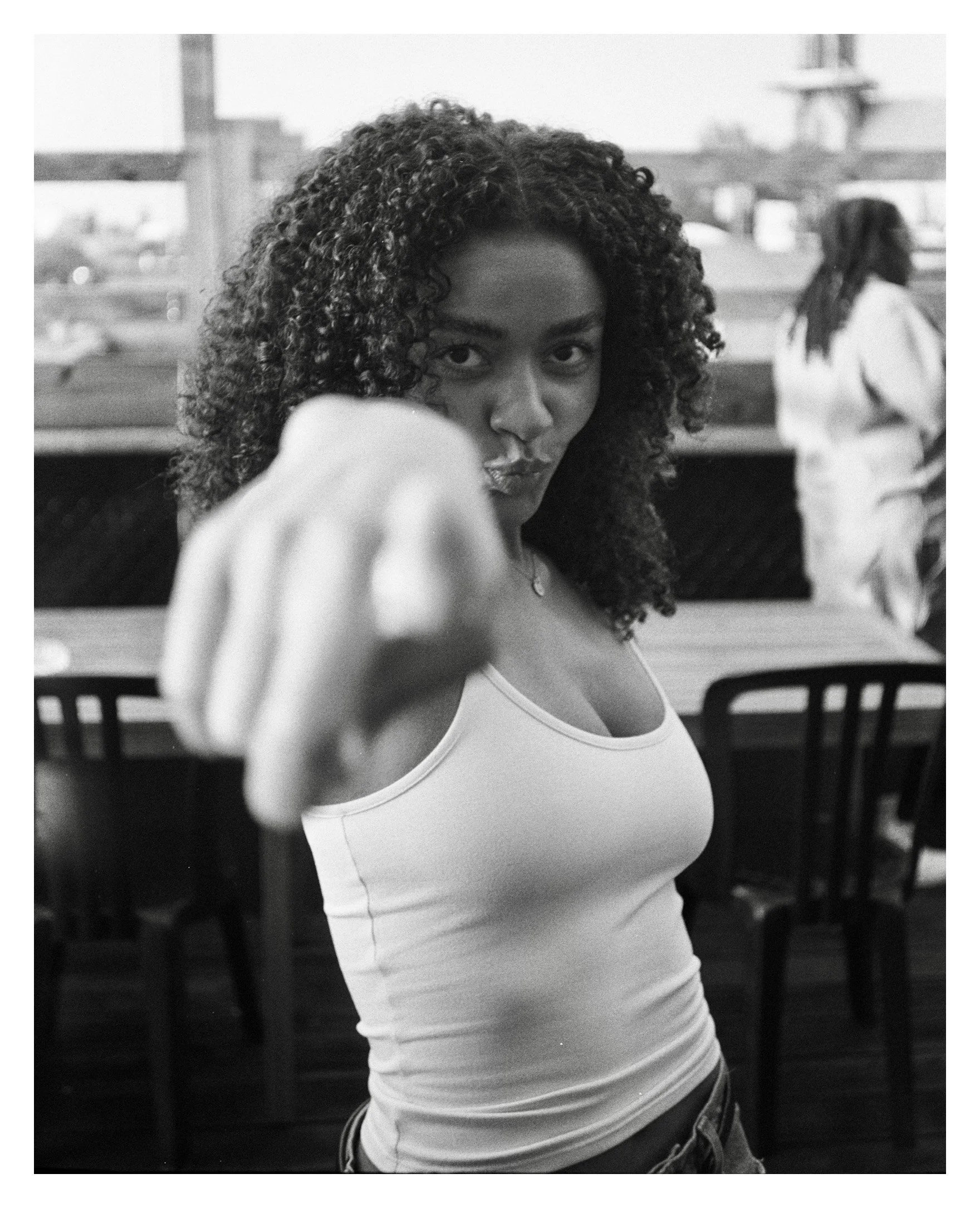 Black and white photo of a woman with curly hair pointing directly at the camera outdoors. She is wearing a sleeveless top and necklace, with a table and person in the background.