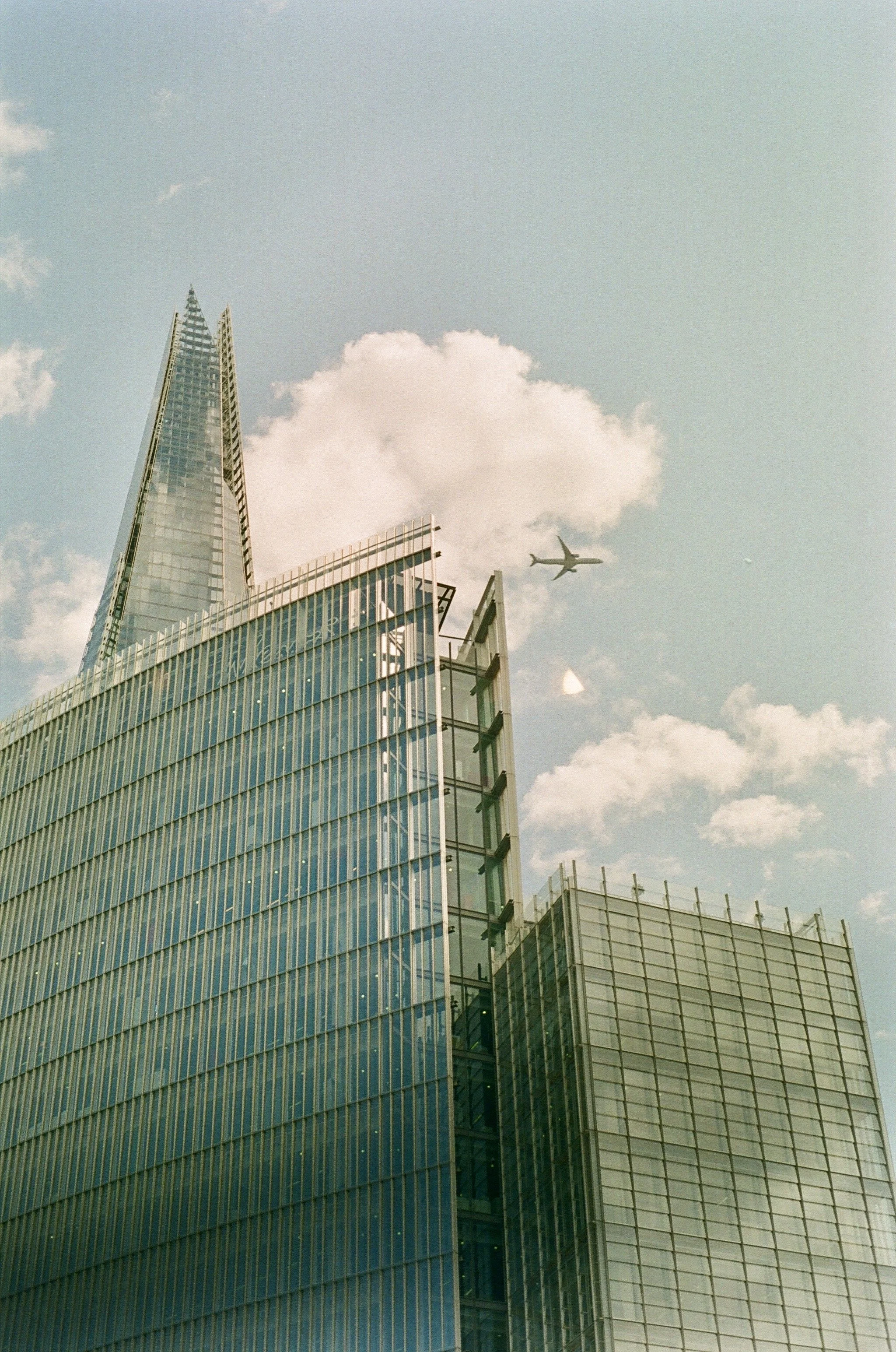 Tall glass skyscraper with modern architecture in a city, with an airplane flying in the sky above and some clouds.