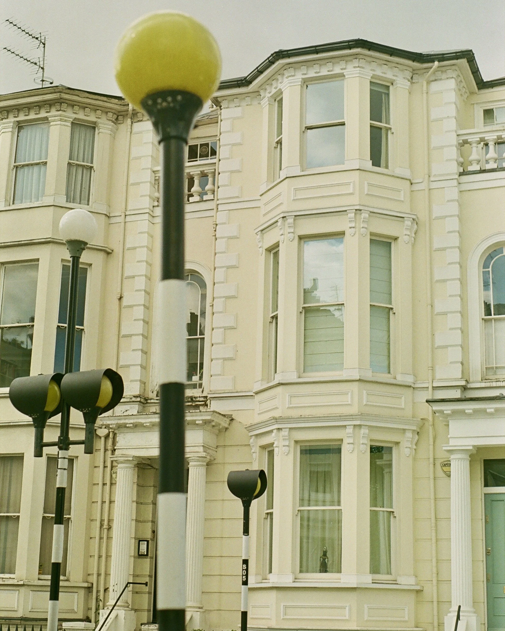 Street view of an ornate cream-colored Victorian building with bay windows and decorative columns, with multiple street lamps and lamps in the foreground.