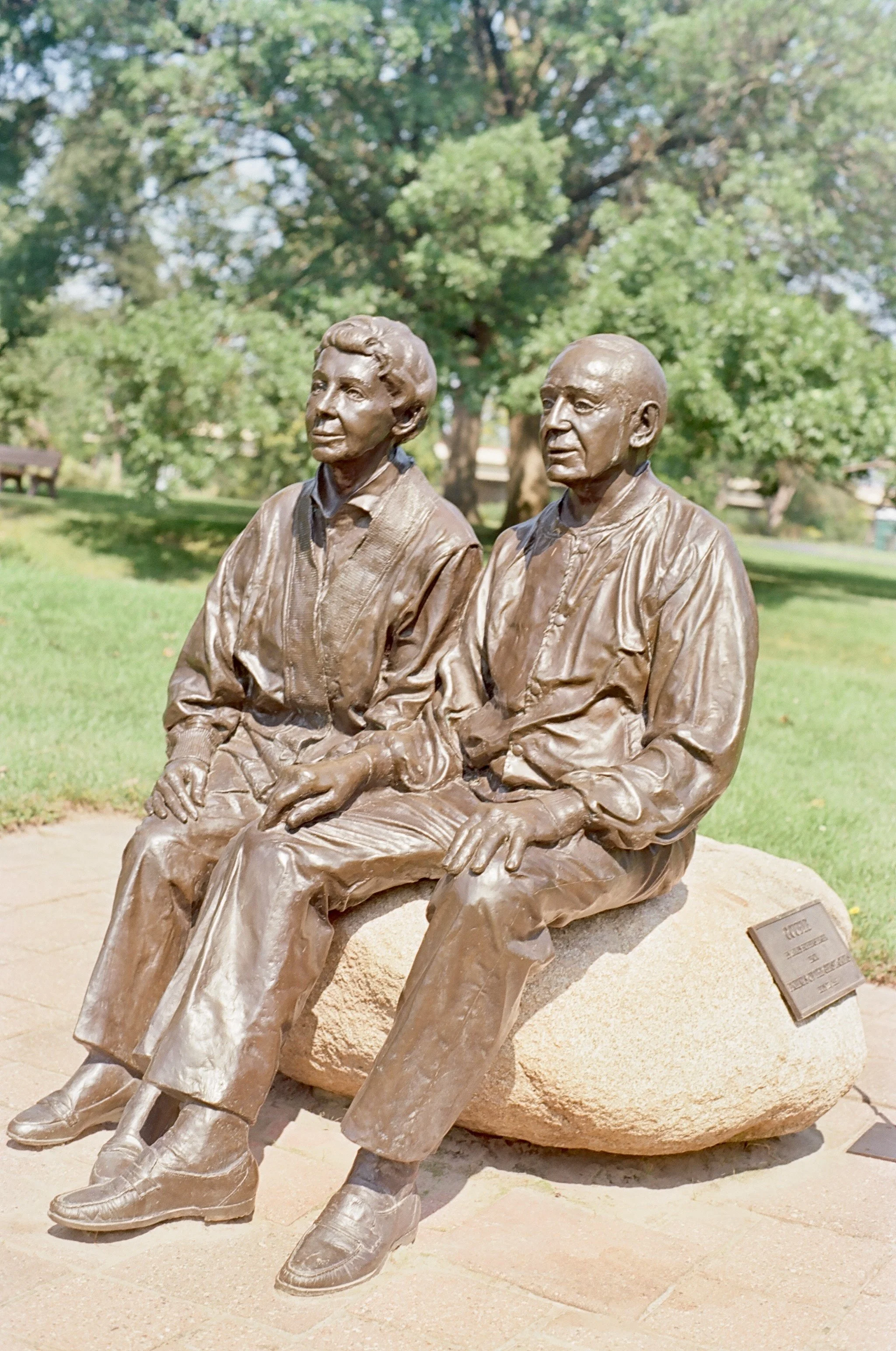 Bronze sculpture of two seated women outdoors in a park, with green trees in the background.