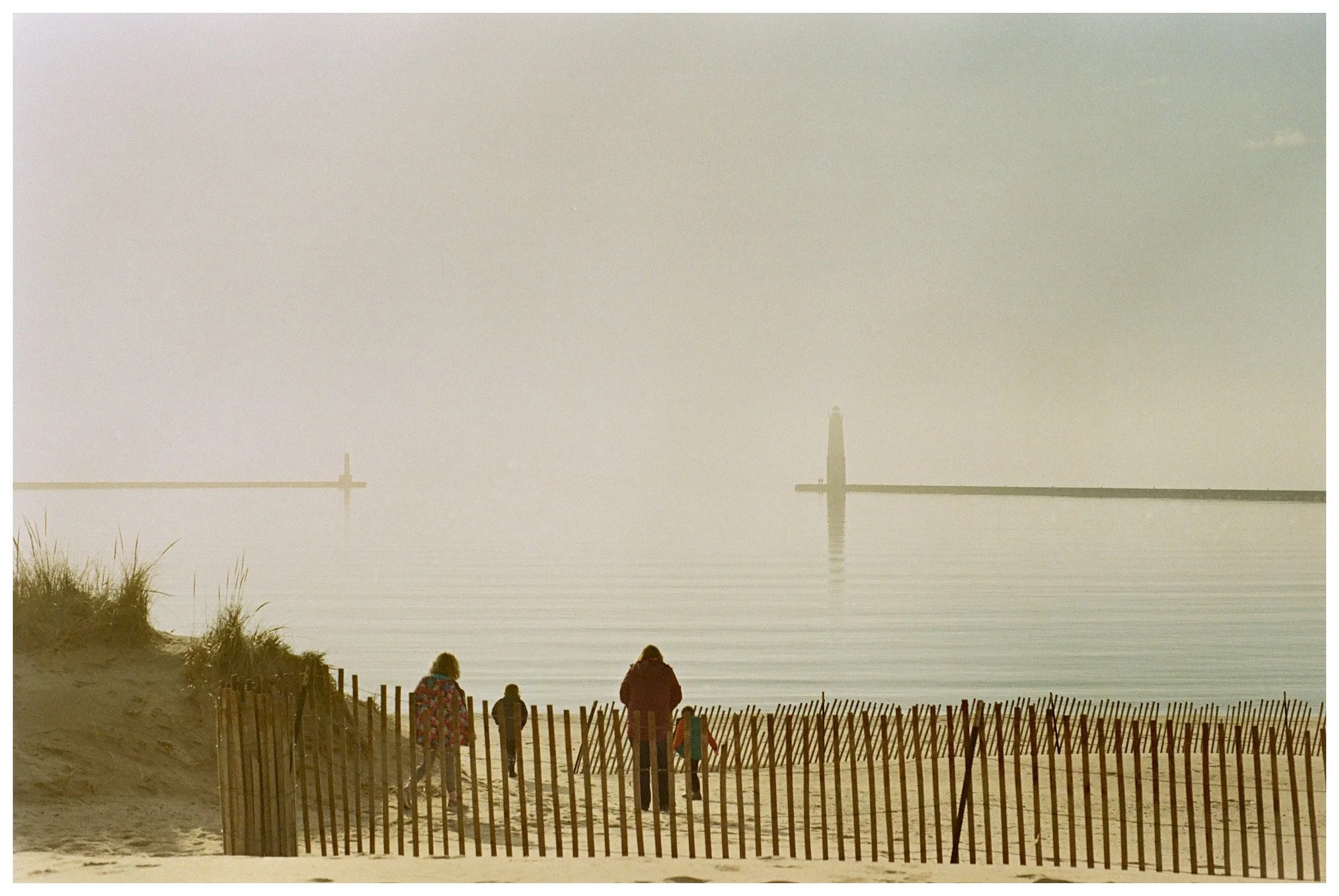 A foggy beach scene with four people walking along the sand near a wooden fence, and two lighthouses in the distance on the water.