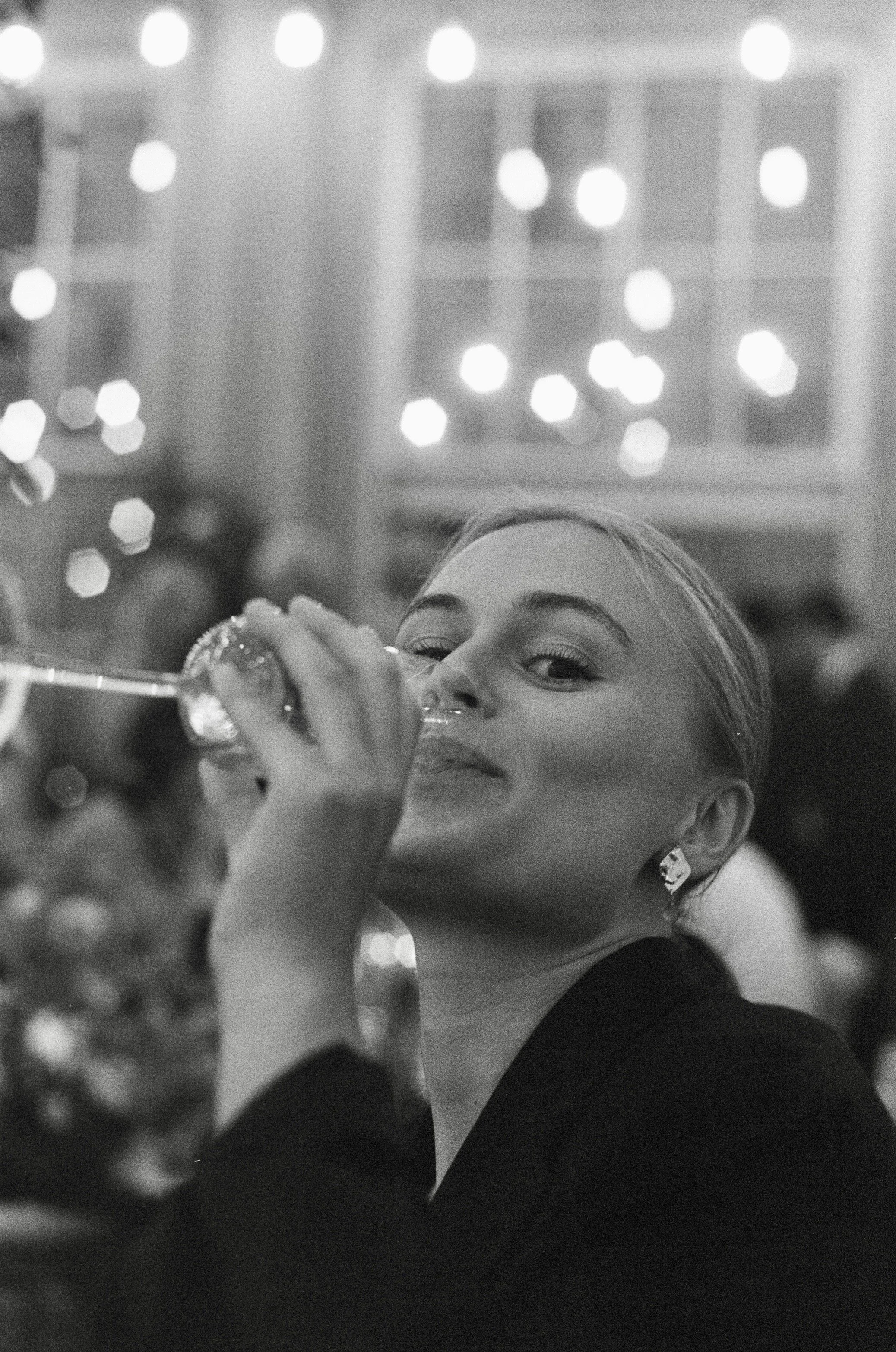 A woman with earrings holding a glass of champagne at a festive event, with blurred lights and people in the background.