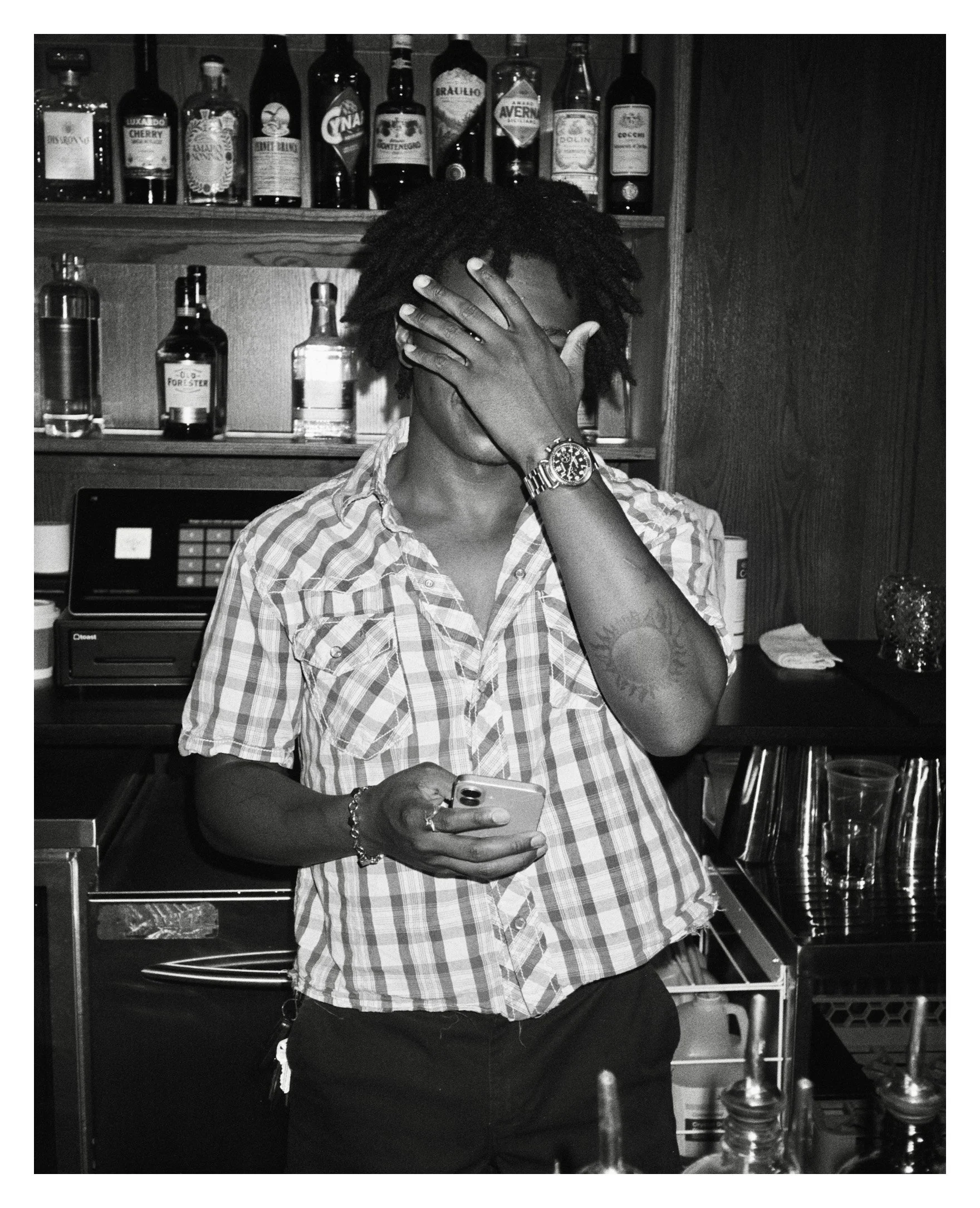 A person with dreadlocks covering their face, wearing a checkered shirt, holds a smartphone in one hand and covers their face with the other hand, standing behind a bar with liquor bottles on the shelves.