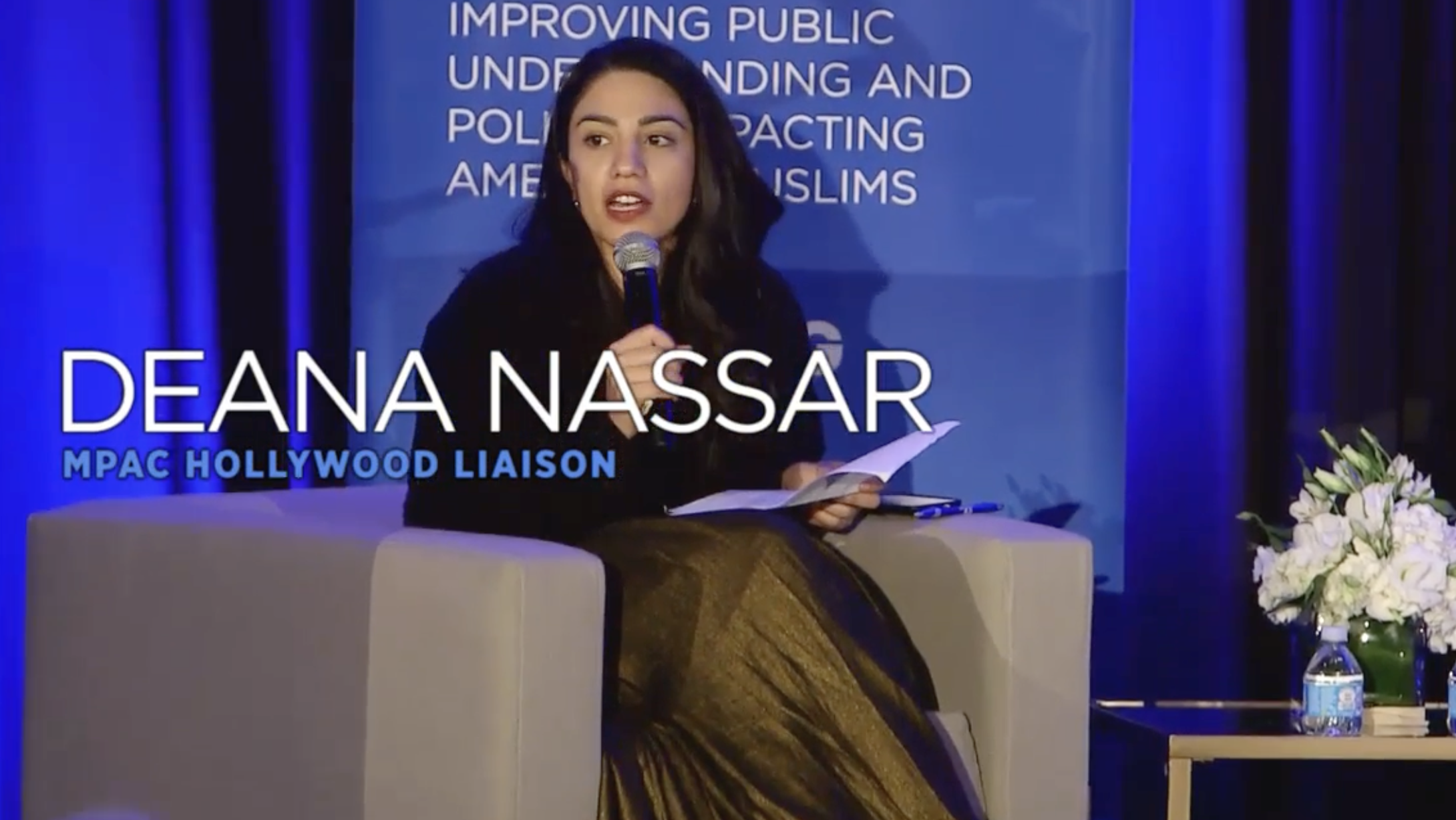 Deana Nassar speaking into a microphone during a panel discussion at an event focused on improving public understanding and impacting Muslims in America, with a blue background featuring white text and a floral arrangement on a side table.
