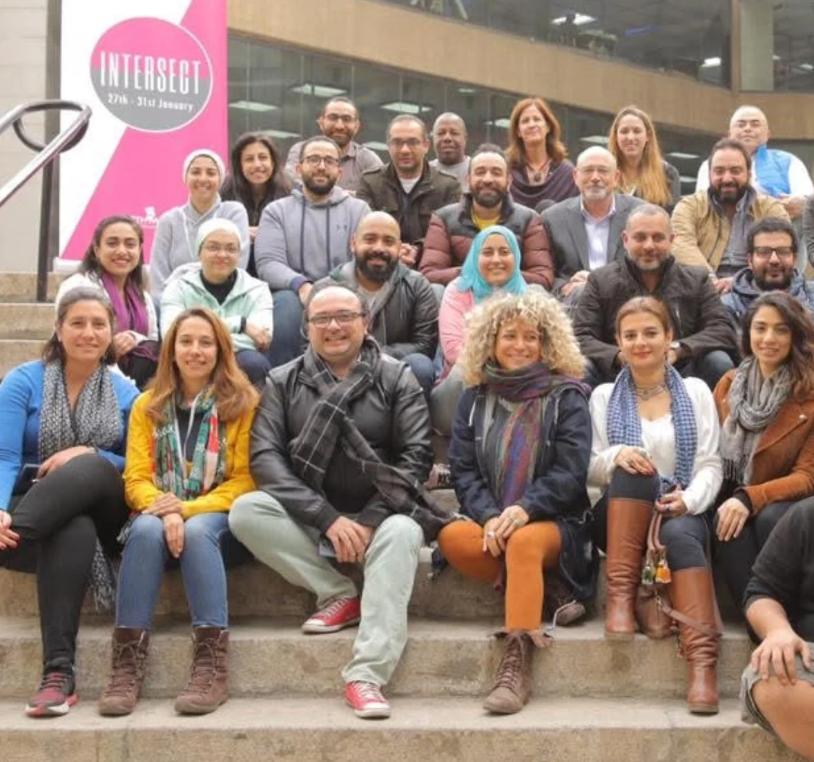 Group of diverse people sitting on steps outside a building with a pink banner that reads "INTERSECT 27th-31st January."