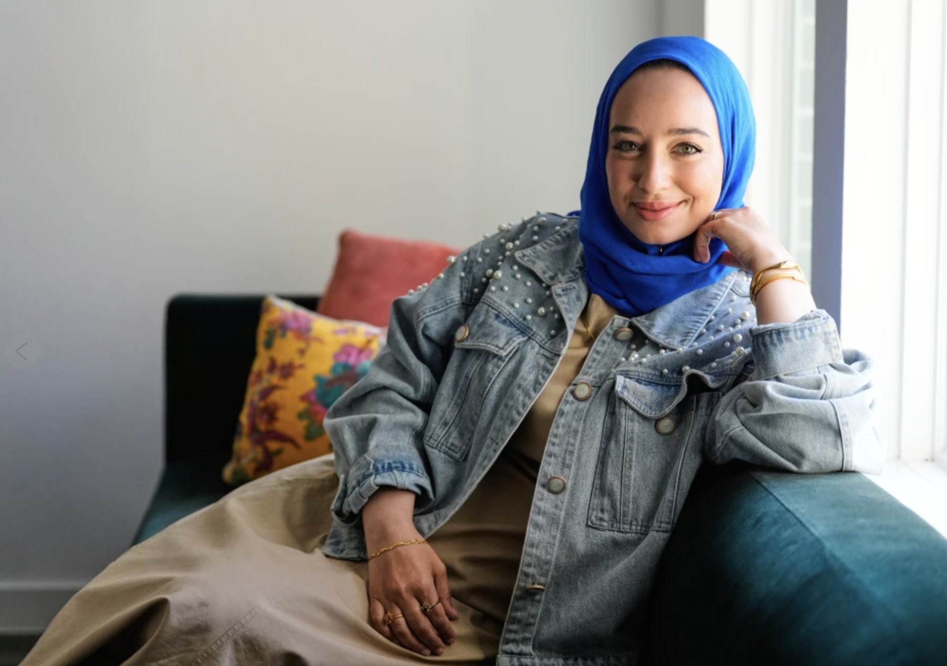 A woman wearing a blue headscarf and a denim jacket with pearl embellishments sitting on a sofa by a window, smiling at the camera.