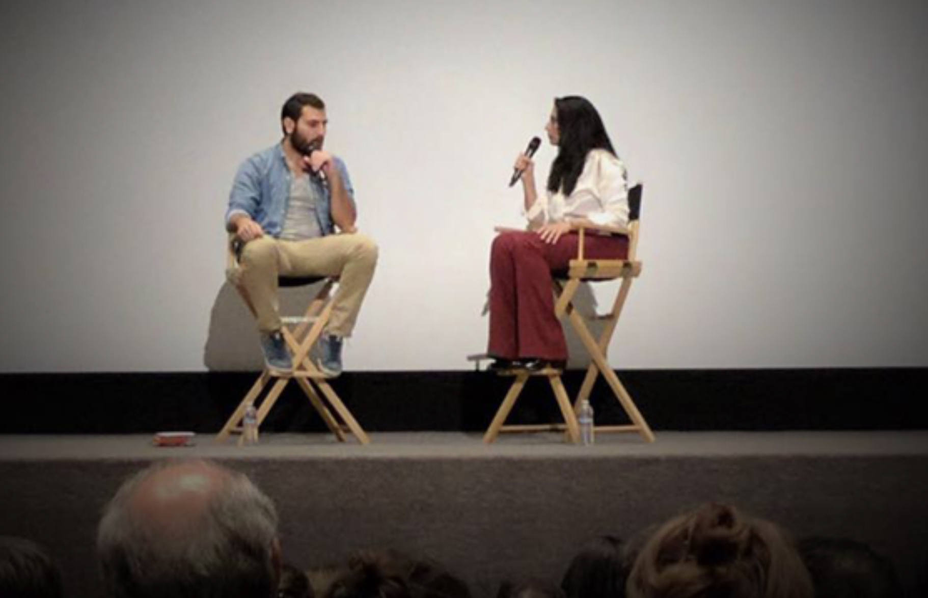 Two people sitting on director's chairs on a stage, engaged in conversation. One is a man with a beard wearing a denim jacket and beige pants, and the other is a woman with long dark hair wearing a white top and red pants. The woman is holding a microphone.