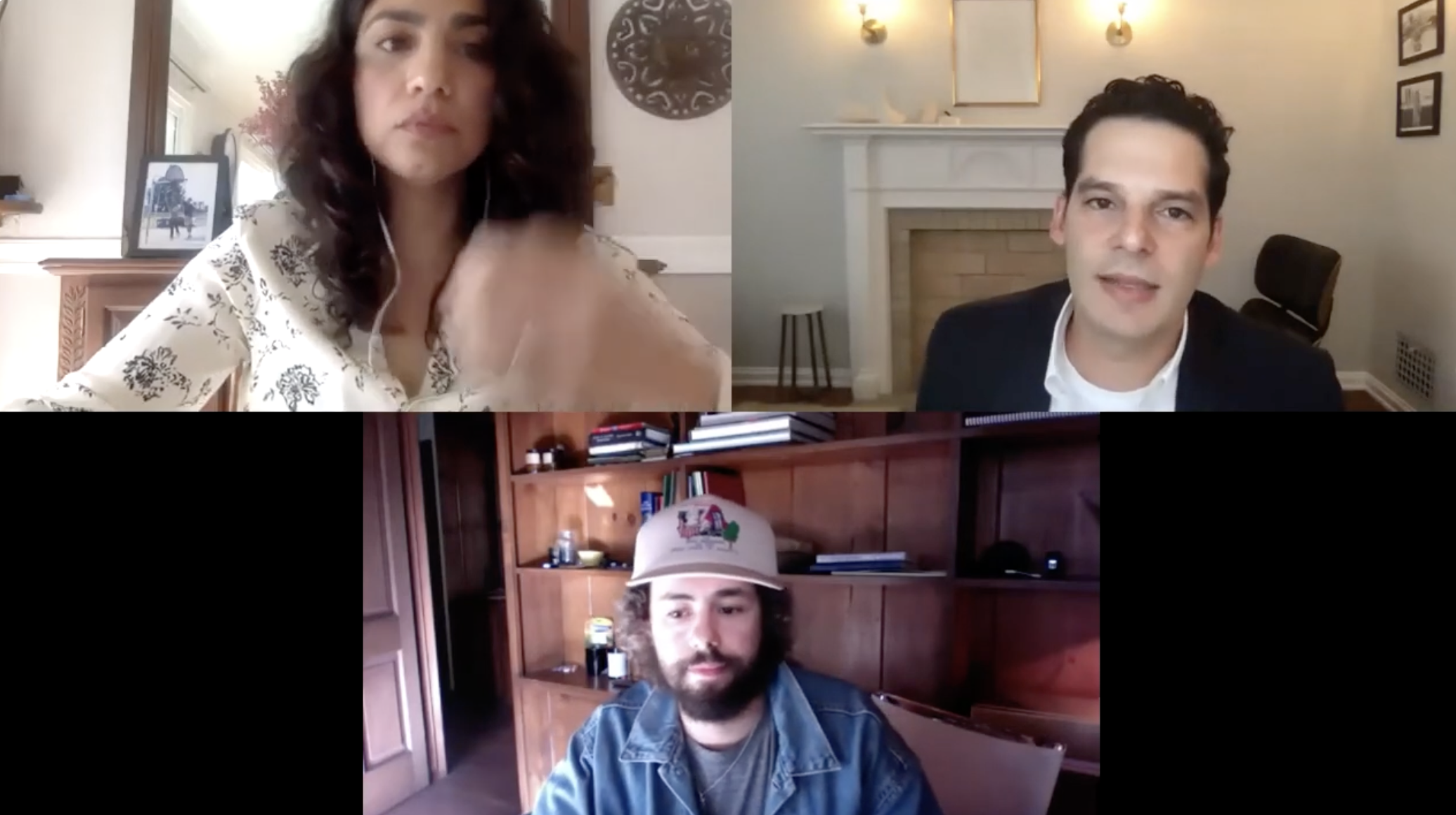 A video call with three participants: a woman with dark wavy hair wearing a patterned shirt in an indoor setting, a man with dark hair and a white shirt and dark blazer in a room with a white fireplace, and a man with a beard and curly hair wearing a blue denim jacket and a cap in a room with wooden shelves.