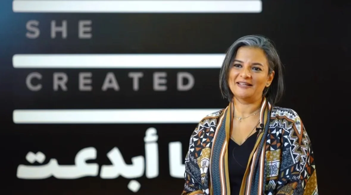 A woman with shoulder-length gray hair wearing a patterned jacket, standing in front of a solid background with the words "SHE CREATED" in large letters.