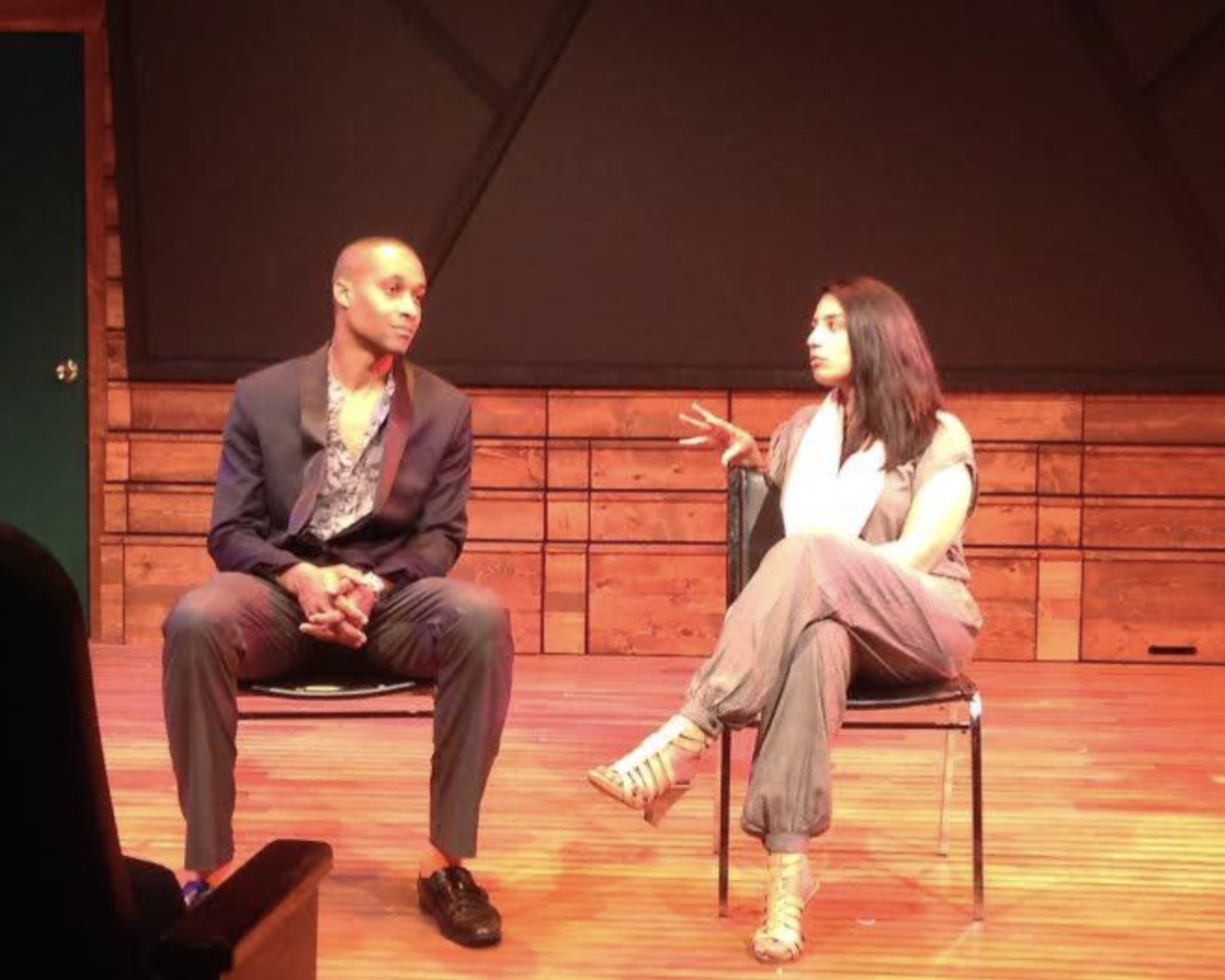 Two people sitting on a stage, engaged in conversation. The man on the left is wearing a dark suit with a patterned shirt, sitting with hands clasped; the woman on the right is wearing a light-colored top and gray pants, gesturing with her hand.