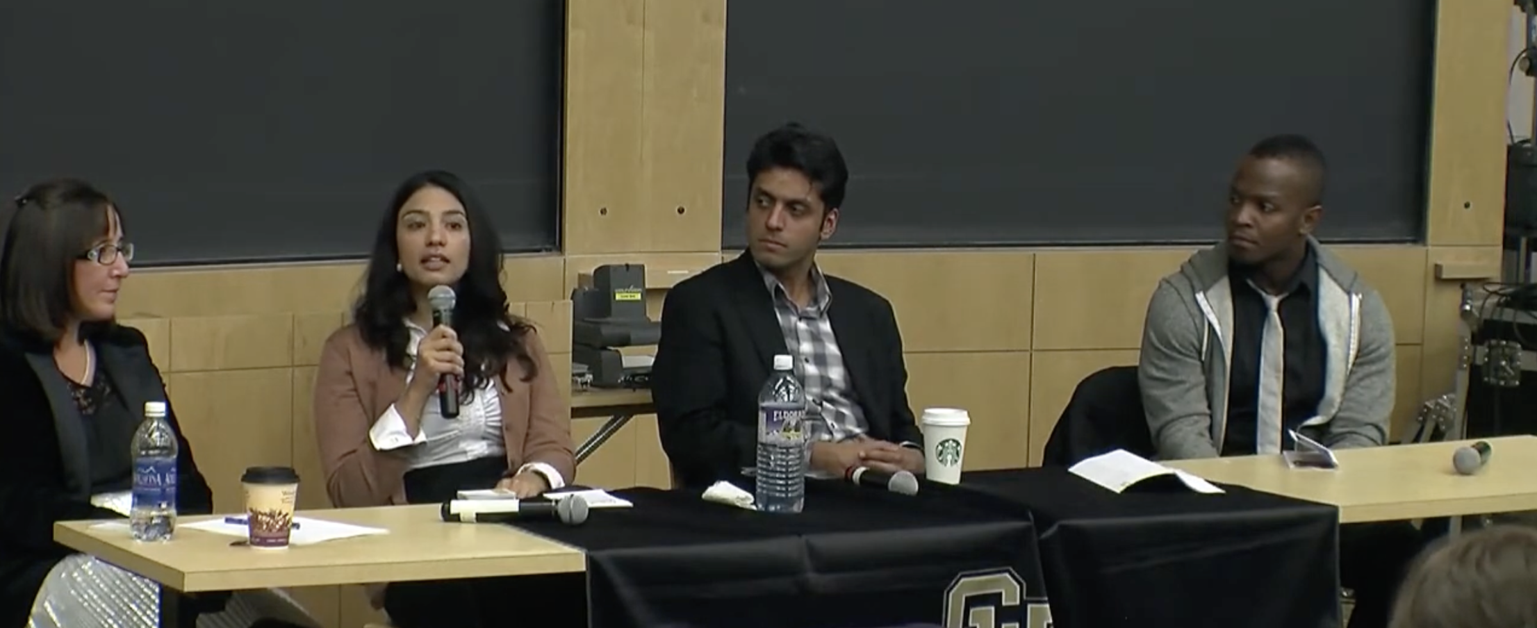 Four panelists sitting at a table during a panel discussion or conference. One woman is speaking into a microphone, and there are water bottles, coffee cups, and notes in front of them.