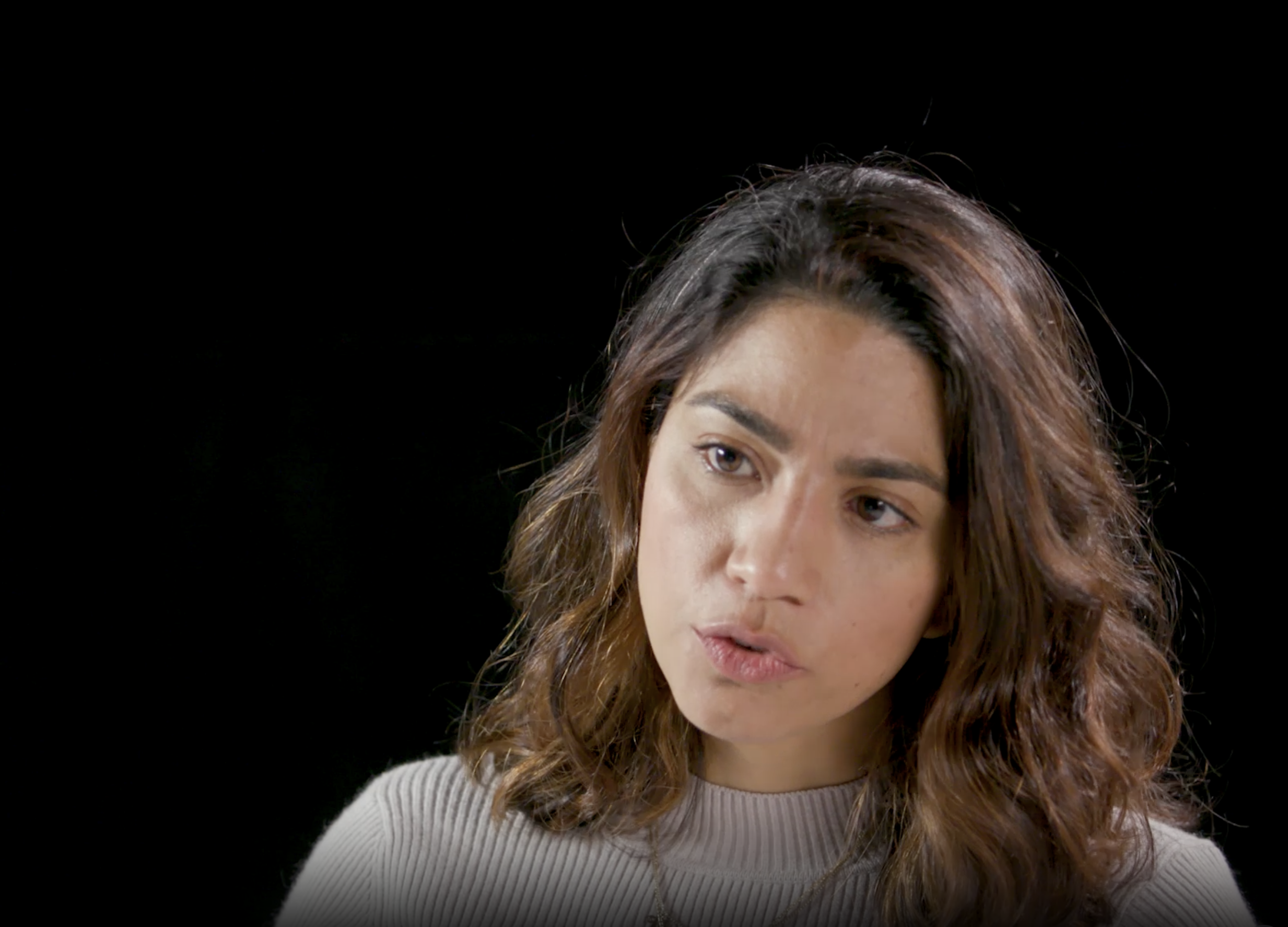 A young woman with wavy brown hair and light skin looks to the side against a black background.