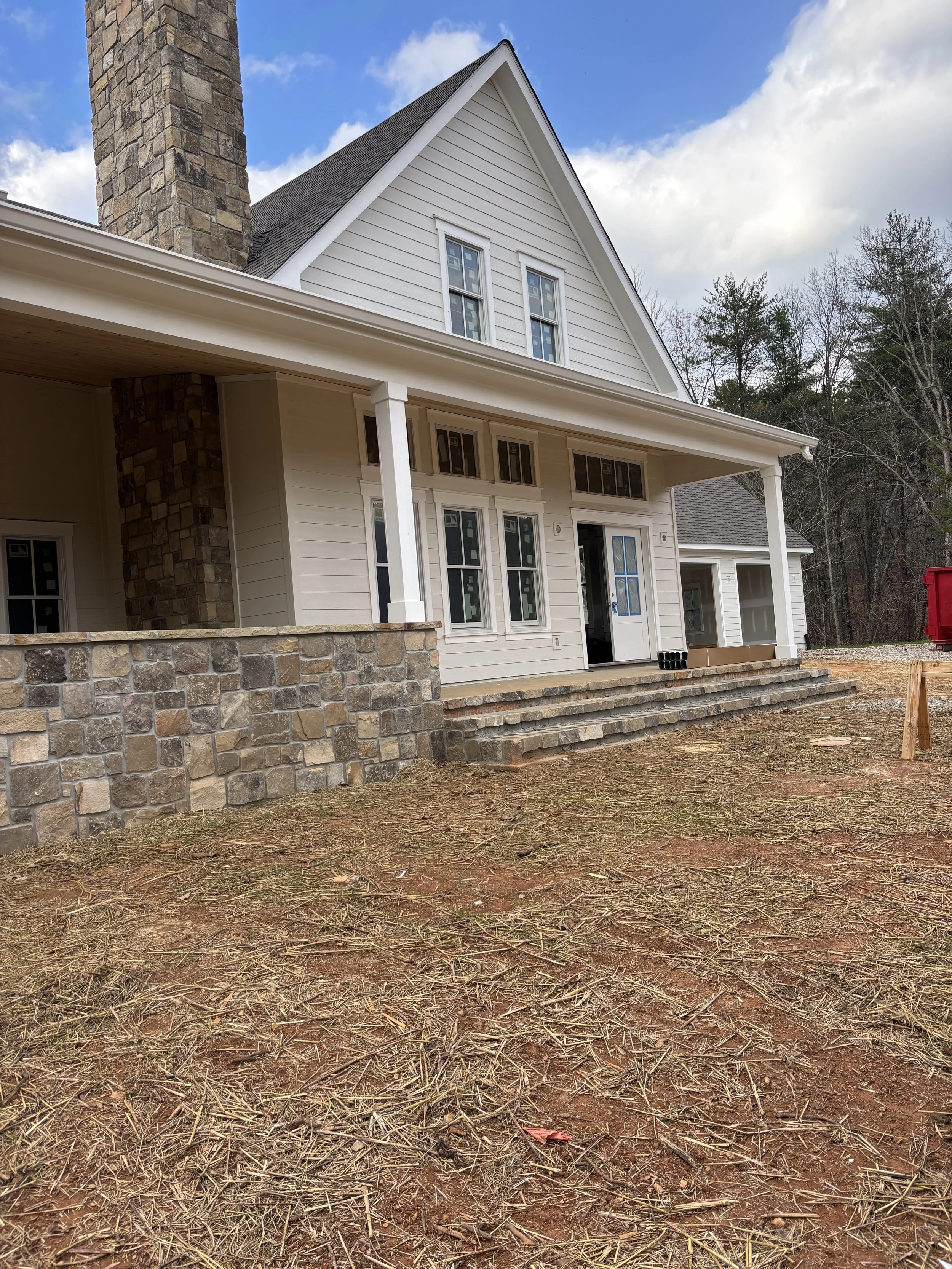 Newly constructed house with a porch, white siding, stone accents, and multiple windows, with steps leading up to the porch and a partially cloudy sky in the background.