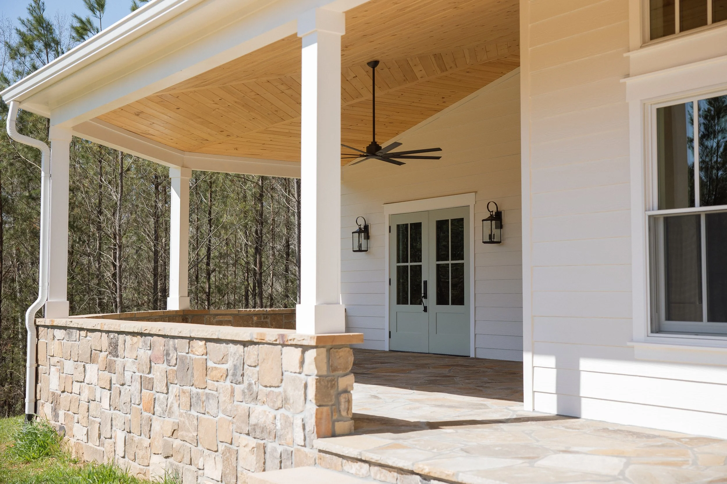 Porch with stone railing, ceiling fan, double doors, and outdoor lighting fixtures, attached to a white house with a wooded background.